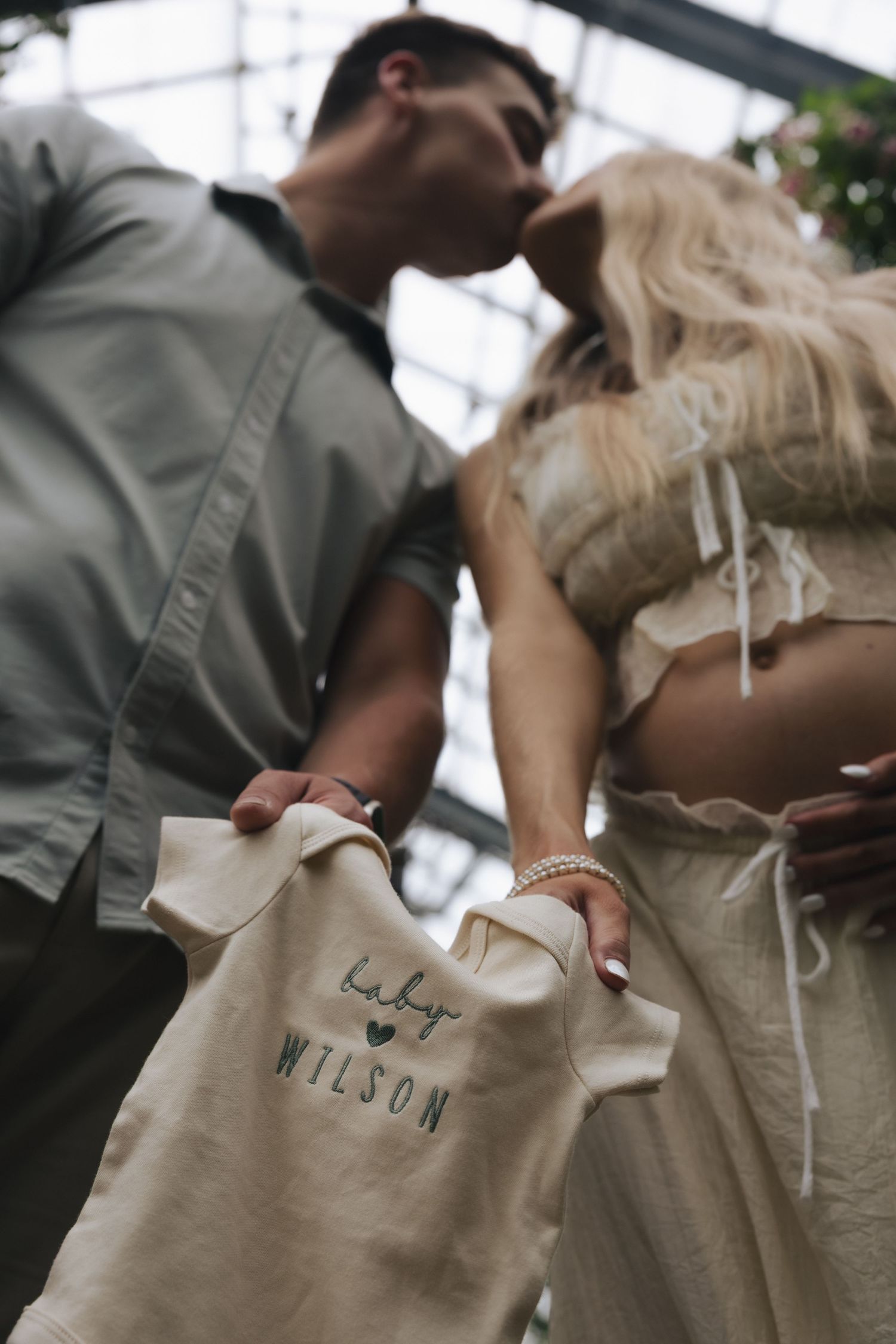 Couple kissing while holding a baby onesie that reads Baby Wilson during a maternity announcement photoshoot.
