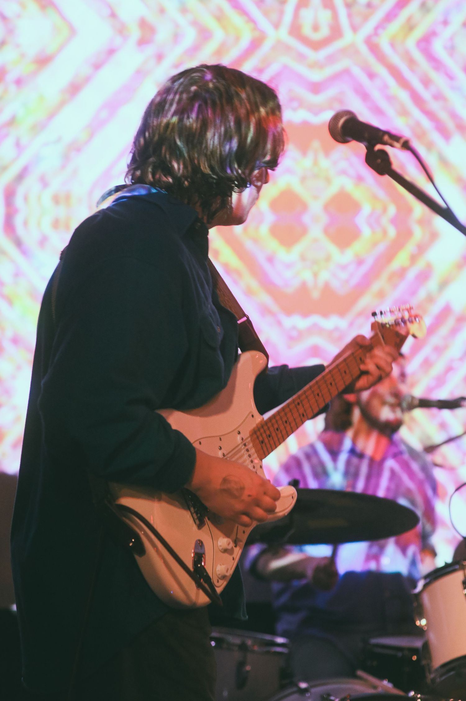 A guitarist plays an electric guitar on stage with psychedelic colorful patterns projected in the background.