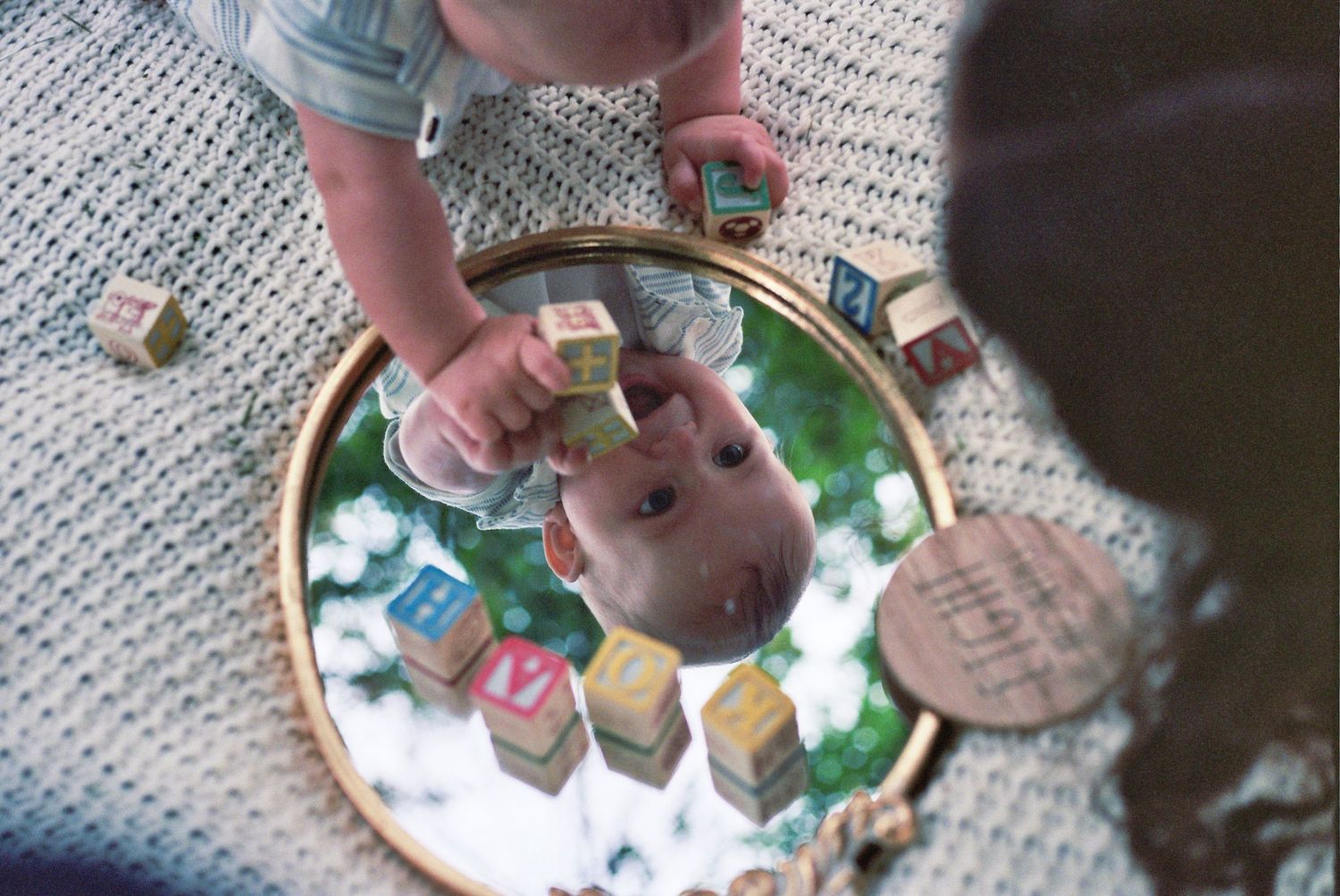 Baby playing with alphabet blocks reflected in a round mirror on a knit blanket.