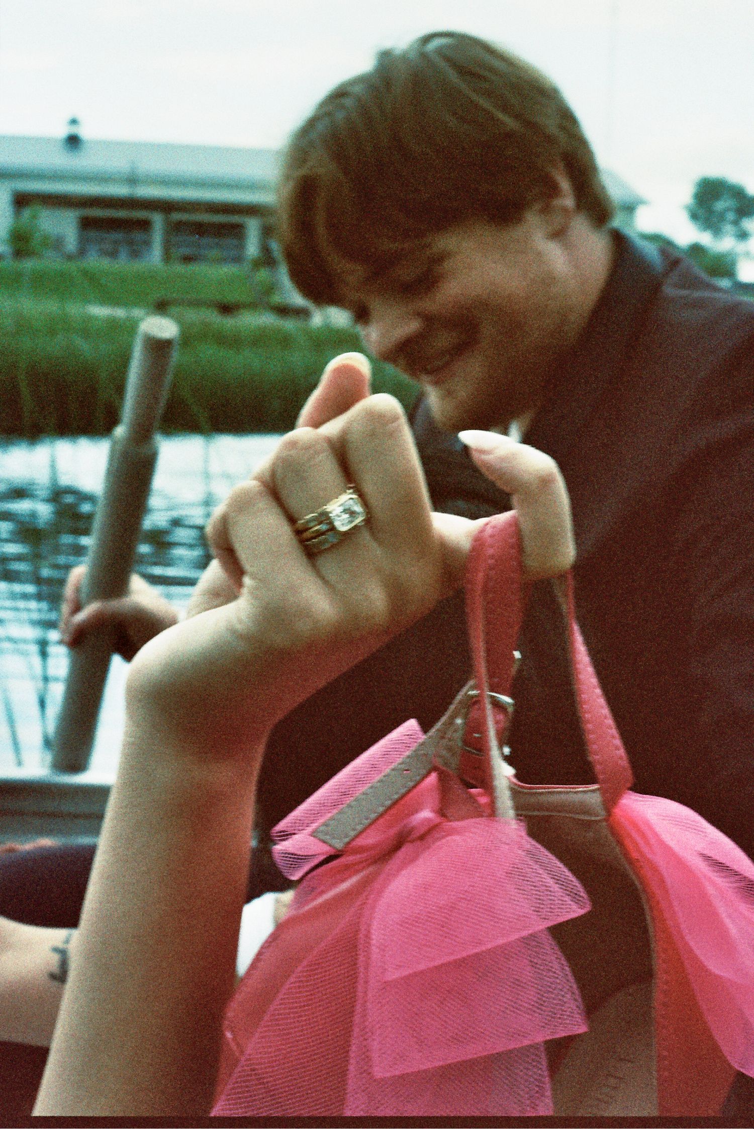 Someone displaying a ring next to a pink tulle fabric detail by a waterfront dock on a vintage-style photograph.