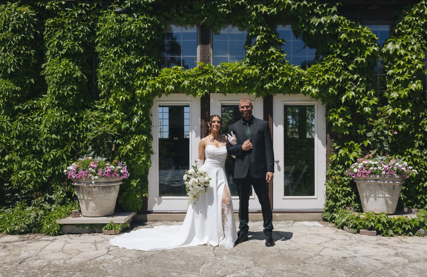 Bride and groom posing in front of ivy-covered venue walls with floral arrangements on sunny wedding day.