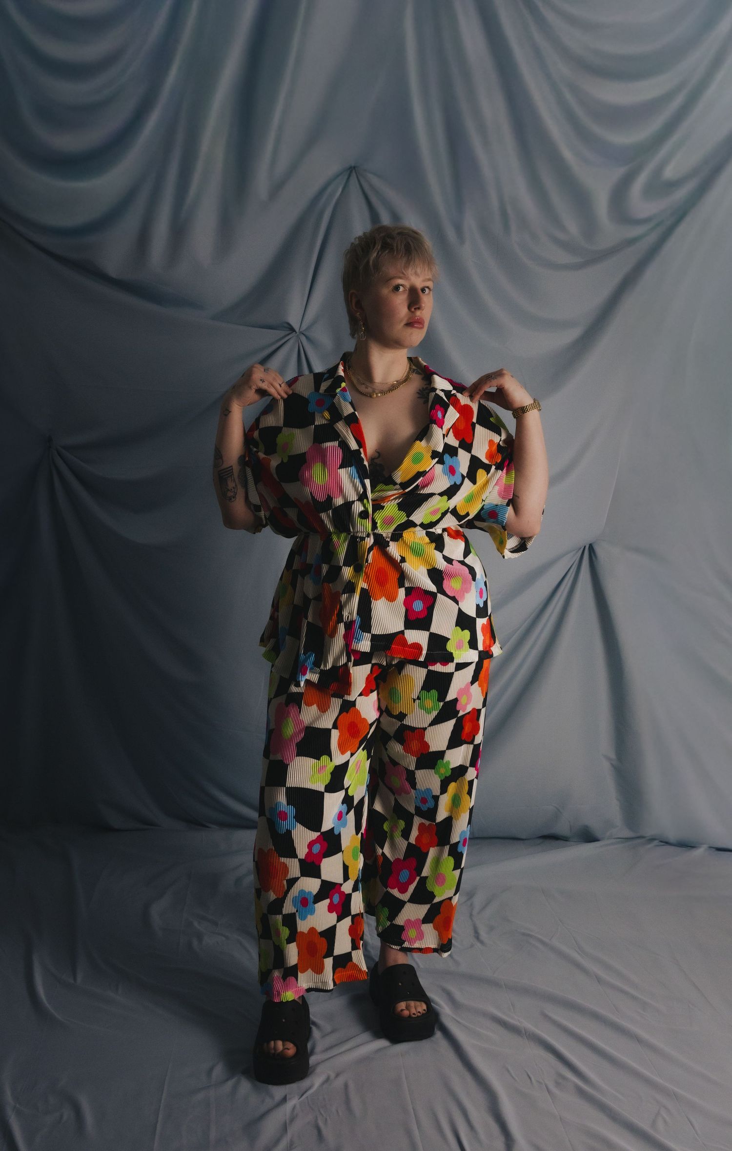 Woman posing in colorful checkered floral jumpsuit with black platform sandals against draped gray backdrop.