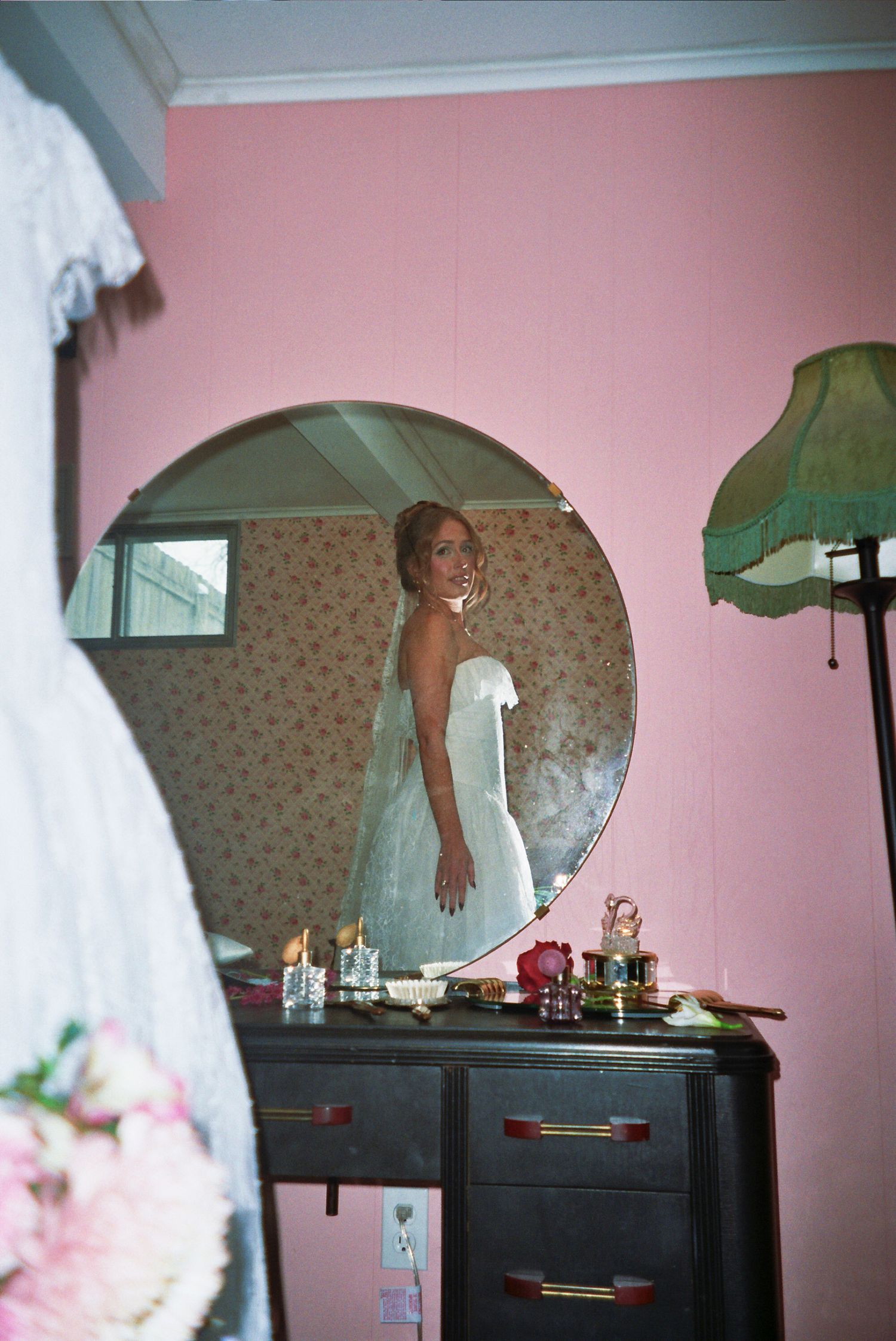 Bride in white wedding dress reflected in round mirror on dark dresser in pink room.