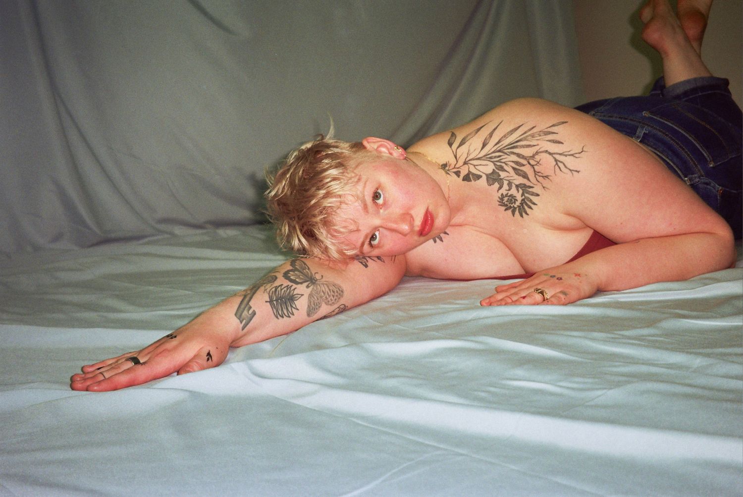 Tattooed person with short blonde hair lying on white sheets, showcasing body art on arms and back.