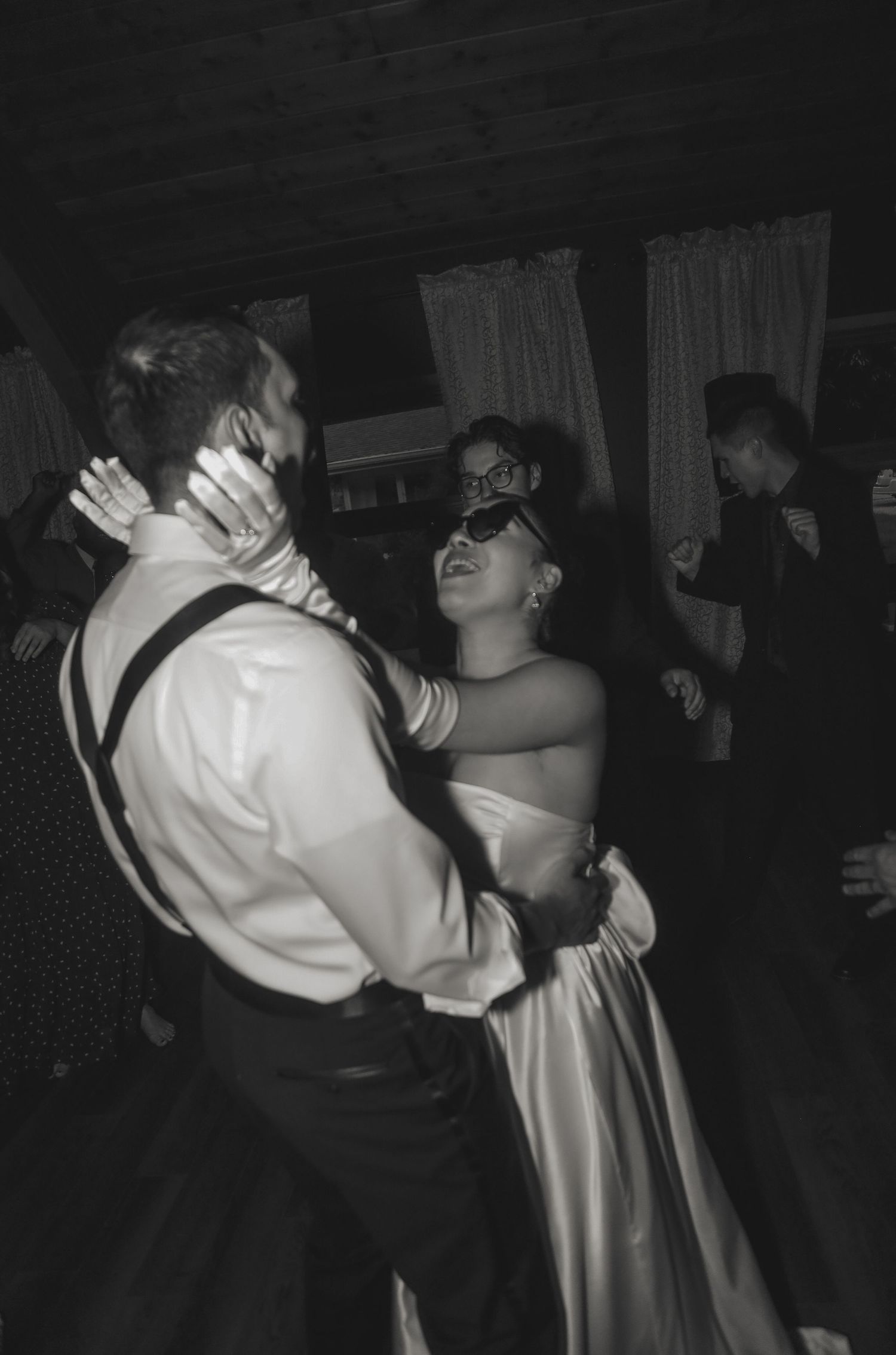 A couple dancing energetically in formal attire at what appears to be a wedding reception, captured in black and white.