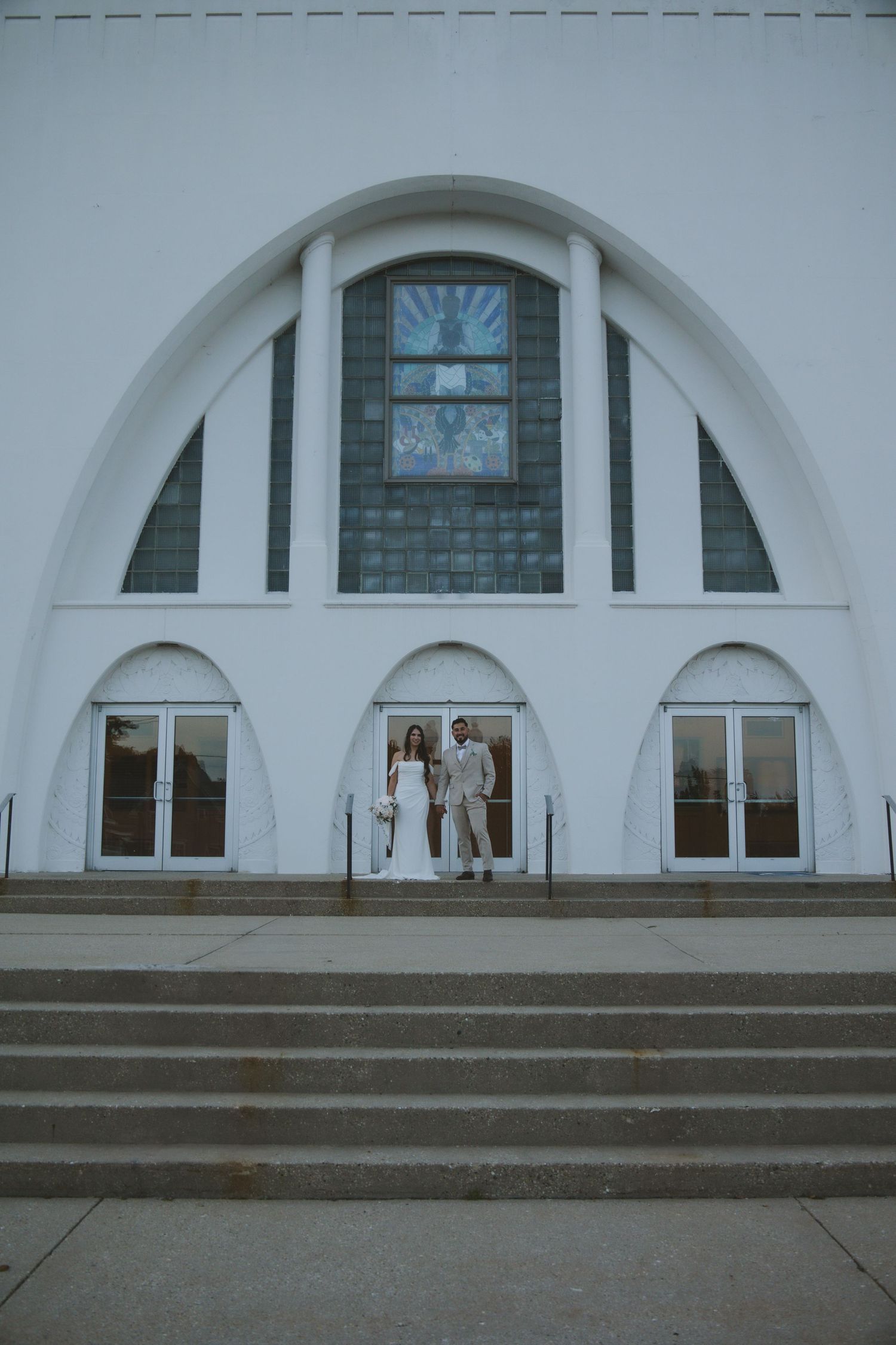 White modern church entrance with arched architecture and stained glass window above stone steps.