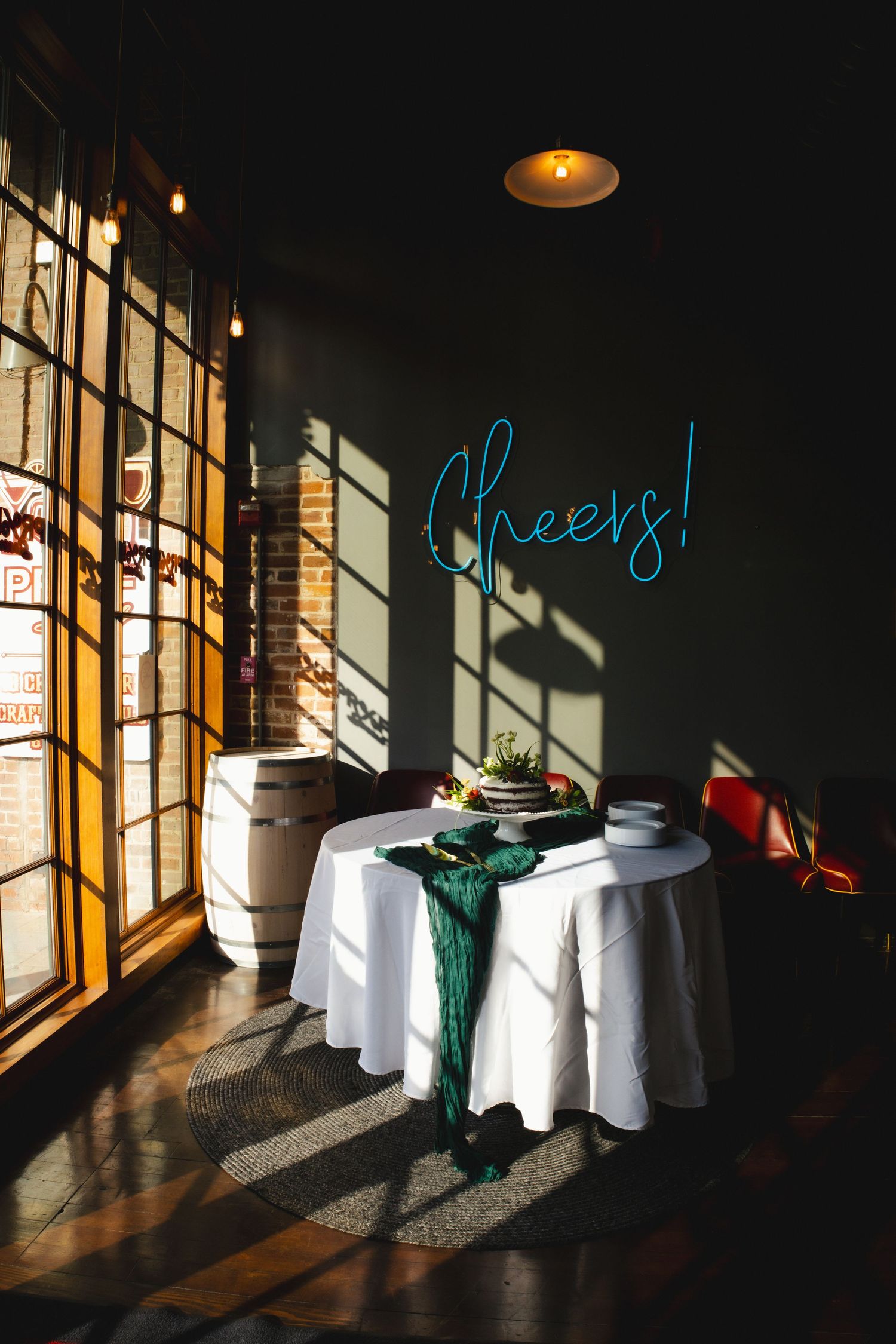 Round white table with neon 'Cheers!' sign and green decor by window casting dramatic shadows in dimly lit room.