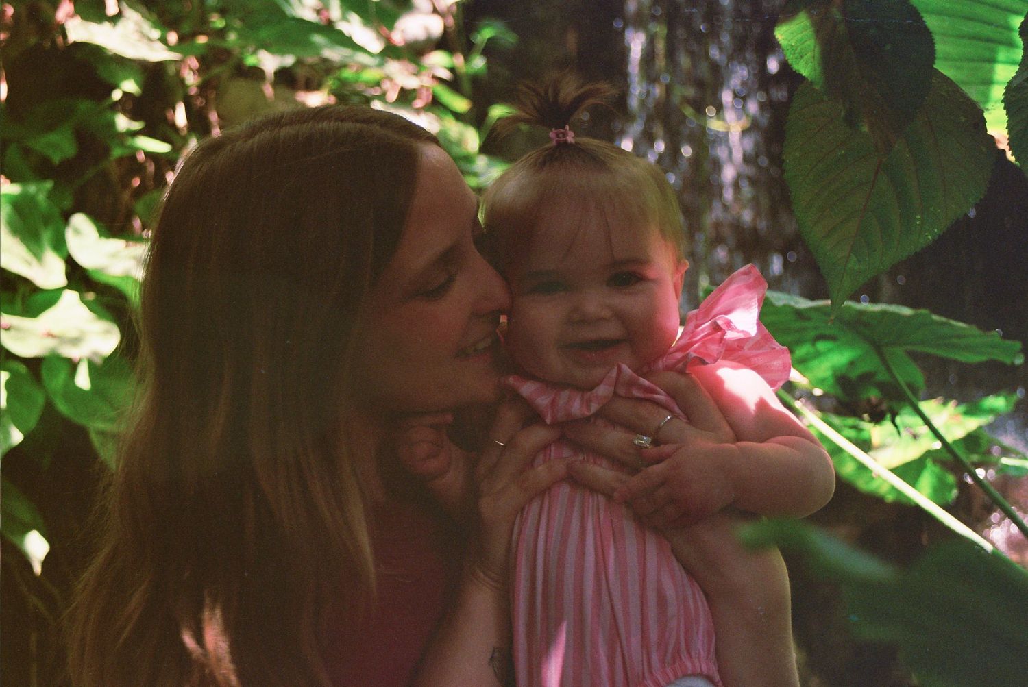 A mother tenderly kisses her smiling baby girl in pink amid lush green tropical foliage.
