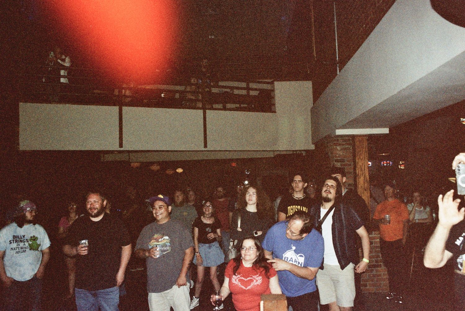 Crowd of people gathered inside a dimly lit venue, watching a live event under red ambient lighting.