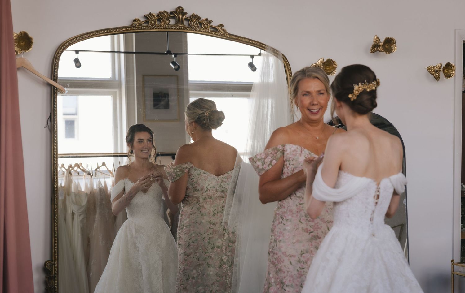 Brides trying on elegant white wedding gowns in a bridal boutique, reflected in a large ornate gold mirror.