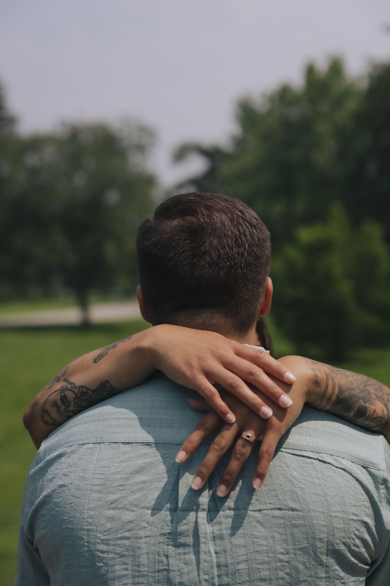 Couple embracing outdoors, man with tattoos wearing blue shirt, woman's hand with ring resting on his shoulder.
