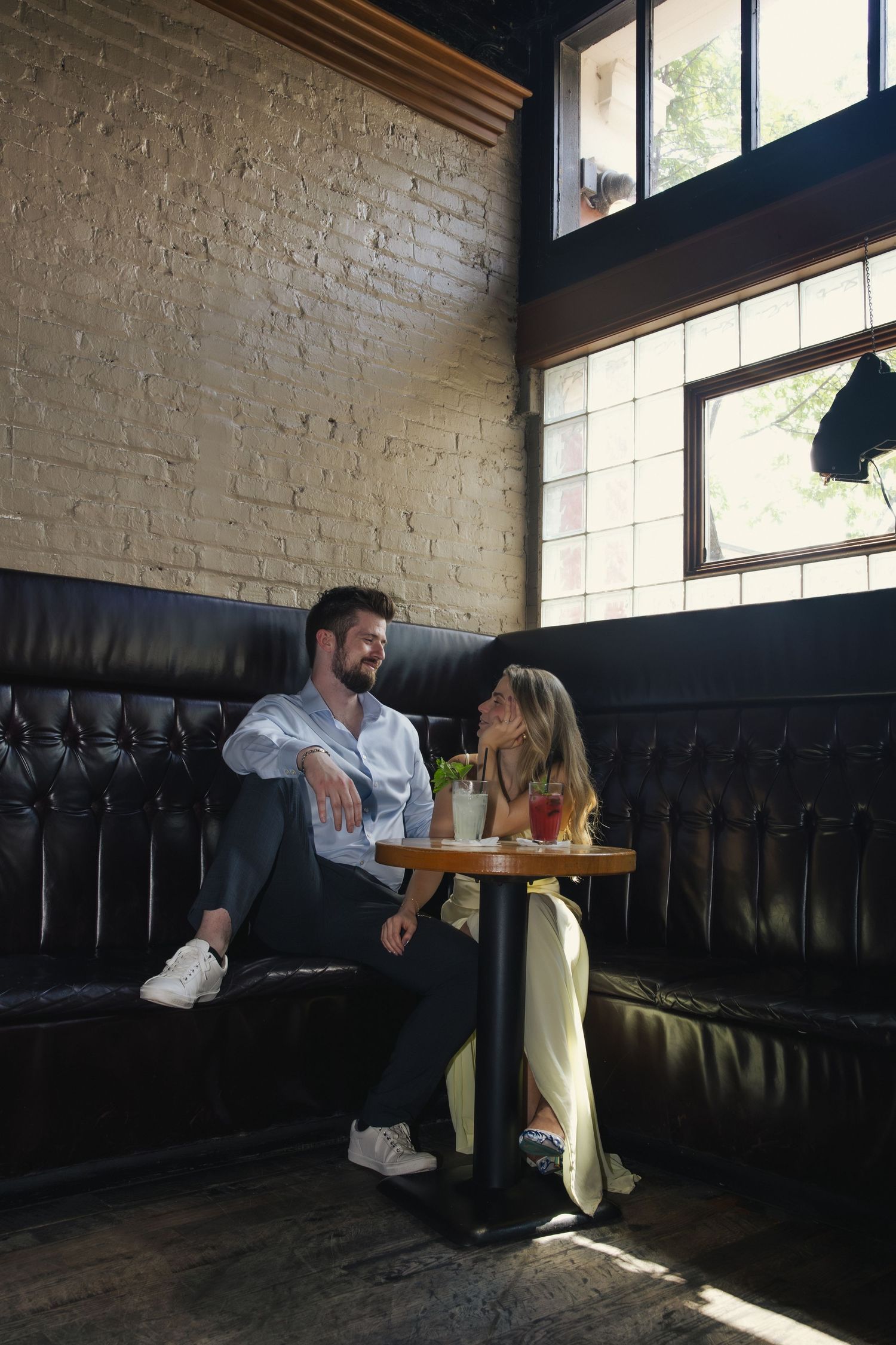 A couple sits together at a small table in a restaurant booth with dark leather seating and white brick walls.