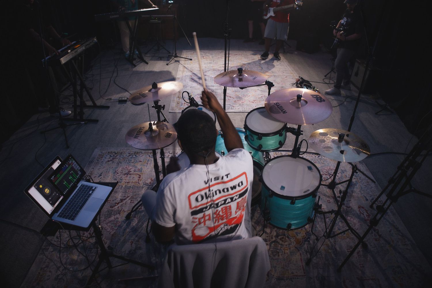 View from behind a drummer performing on a turquoise drum kit in a dark studio setting with musical equipment.