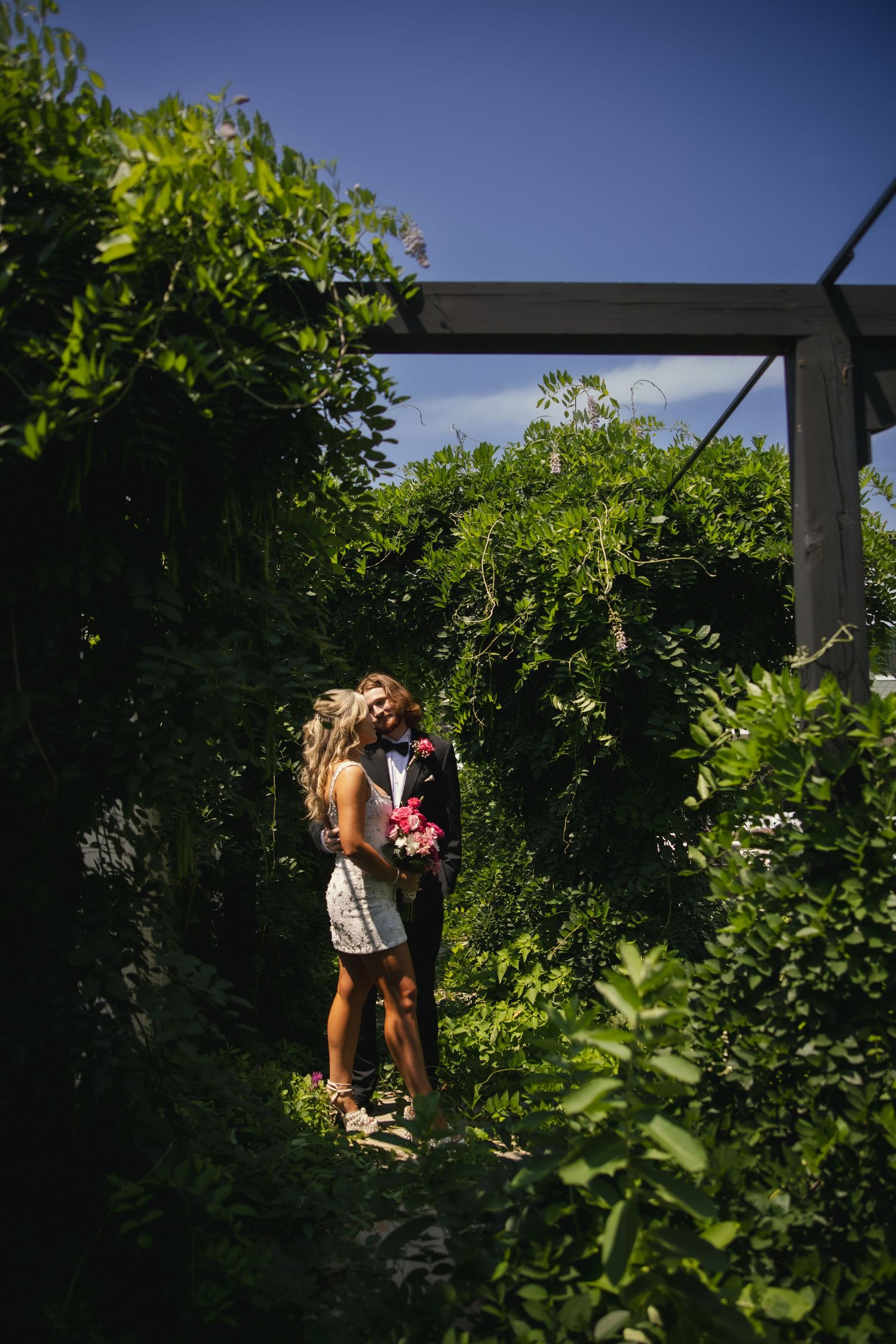 Two people share a tender moment in a lush garden setting with green foliage and bright sunlight.