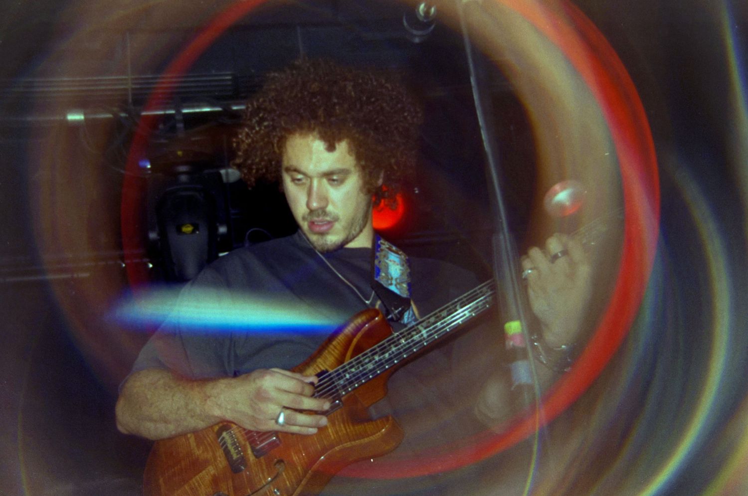 Curly-haired guitarist playing electric guitar on stage, captured through a colorful circular lens effect.