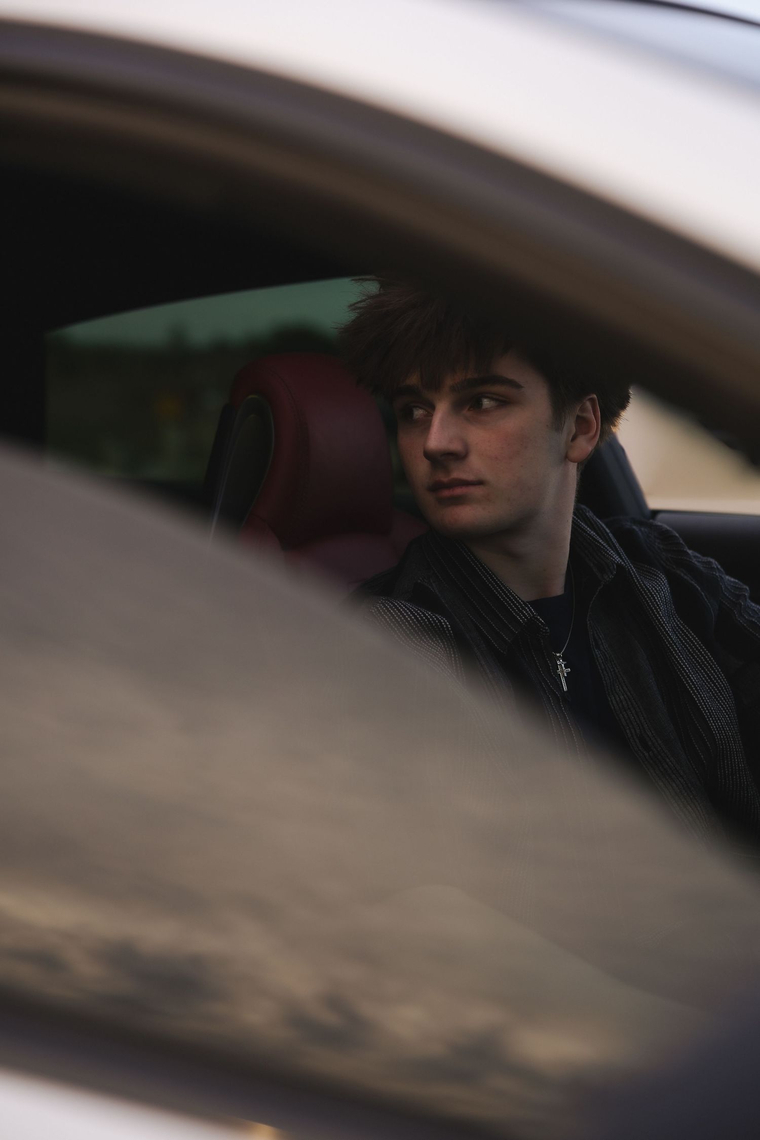 Young man with dark hair sitting in a car with red interior, wearing a dark jacket and cross necklace.