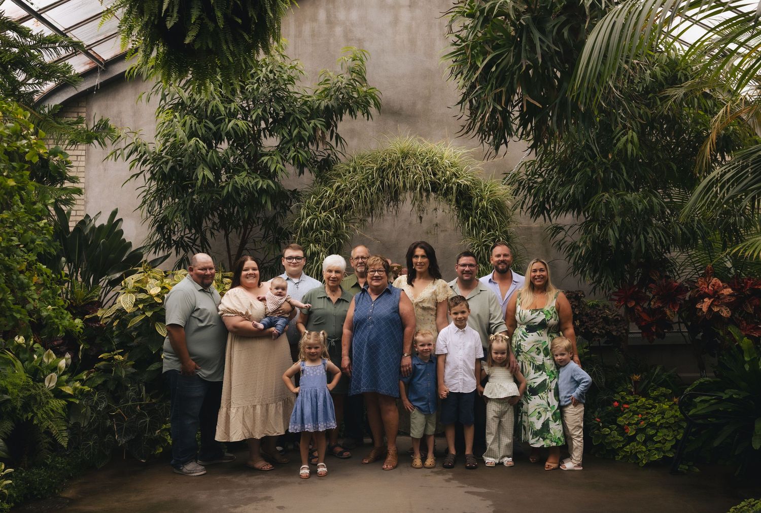 Large family group portrait inside a lush tropical greenhouse with green plants and stone walls.