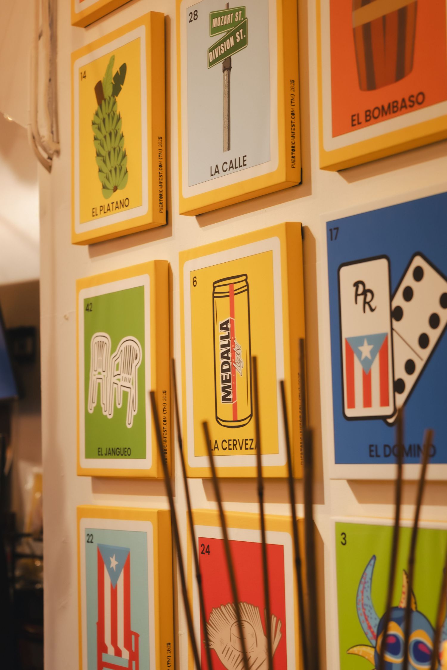 Colorful Puerto Rican loteria cards displayed on a white wall, featuring El Platano, La Calle, Medalla beer, and El Domino.