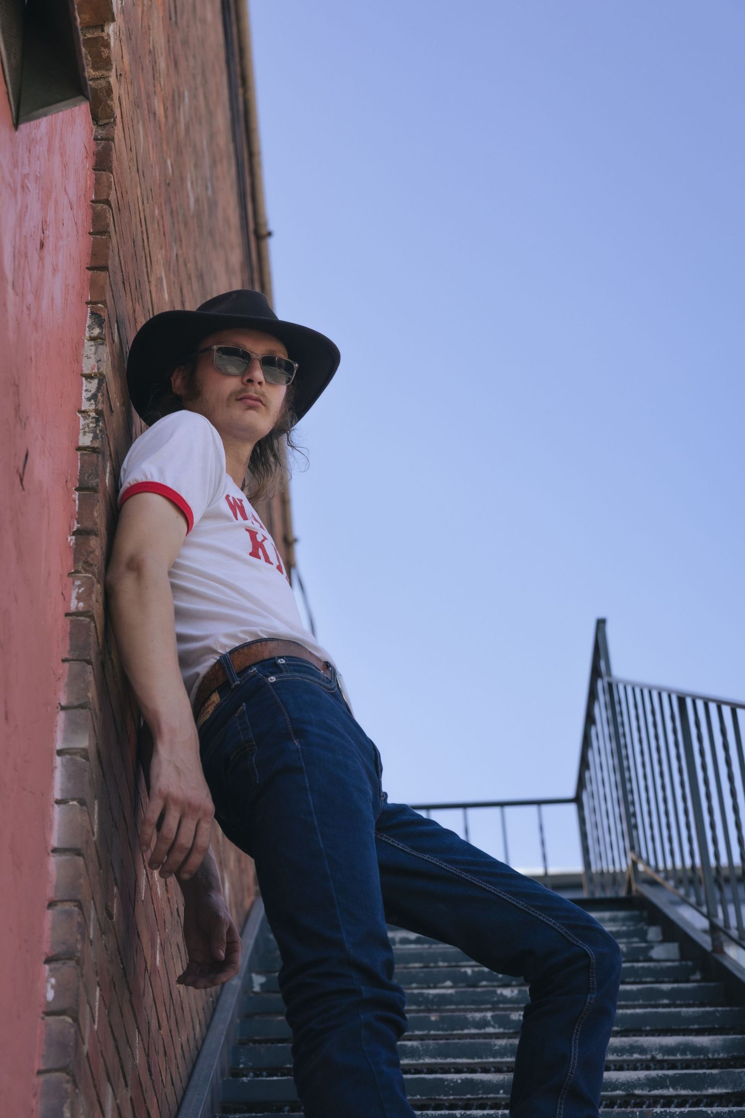 Person in black hat, sunglasses, jeans, and white tee leaning against brick wall near outdoor staircase under blue sky.