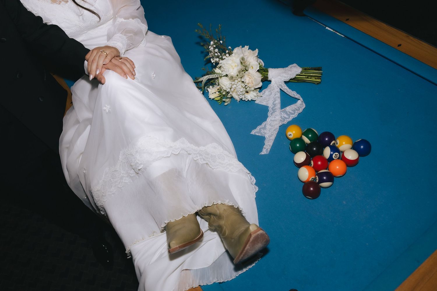 Bride in white wedding dress sitting on blue pool table with billiard balls and floral bouquet nearby.