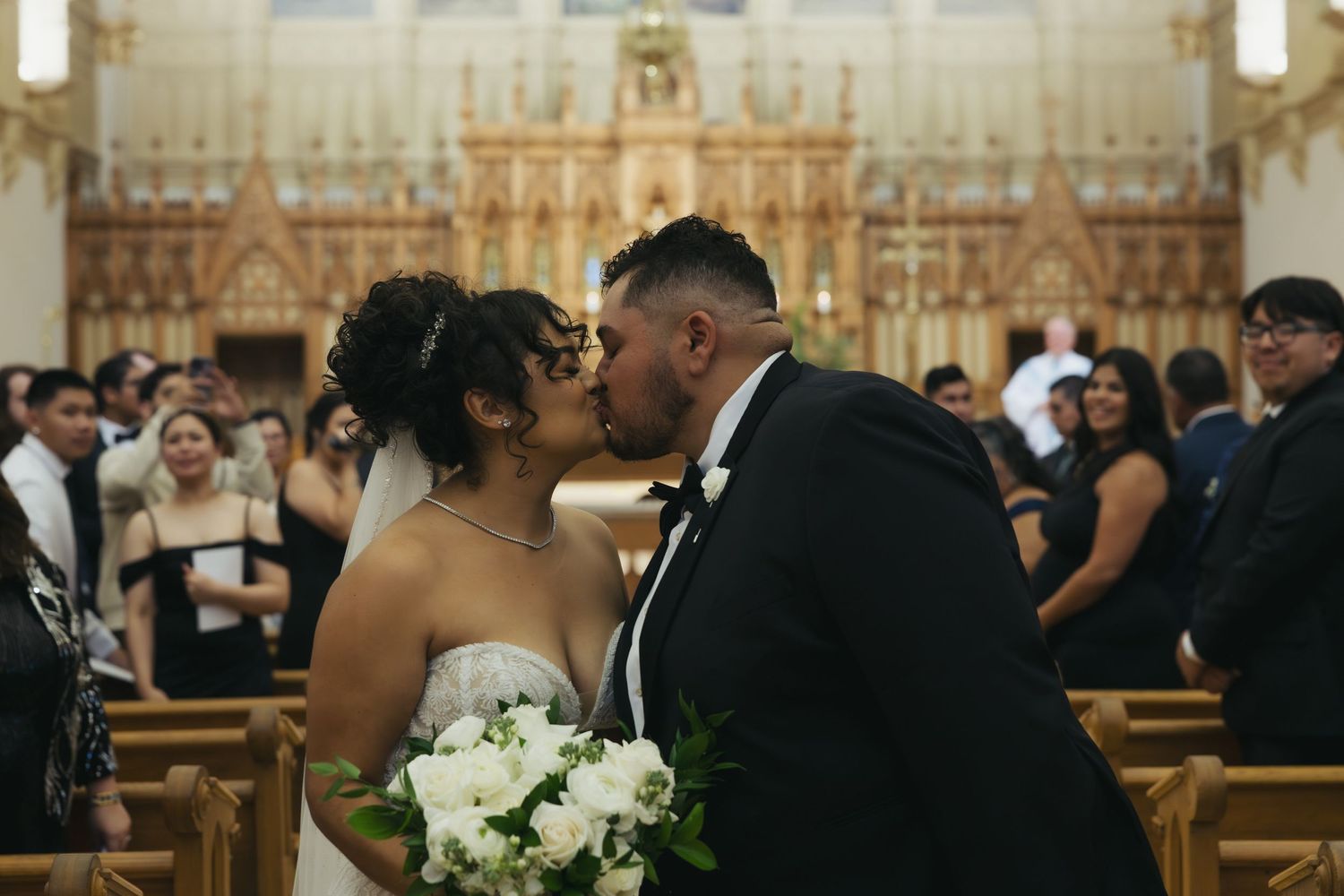 A wedding ceremony takes place inside an ornate church sanctuary with guests seated in wooden pews.