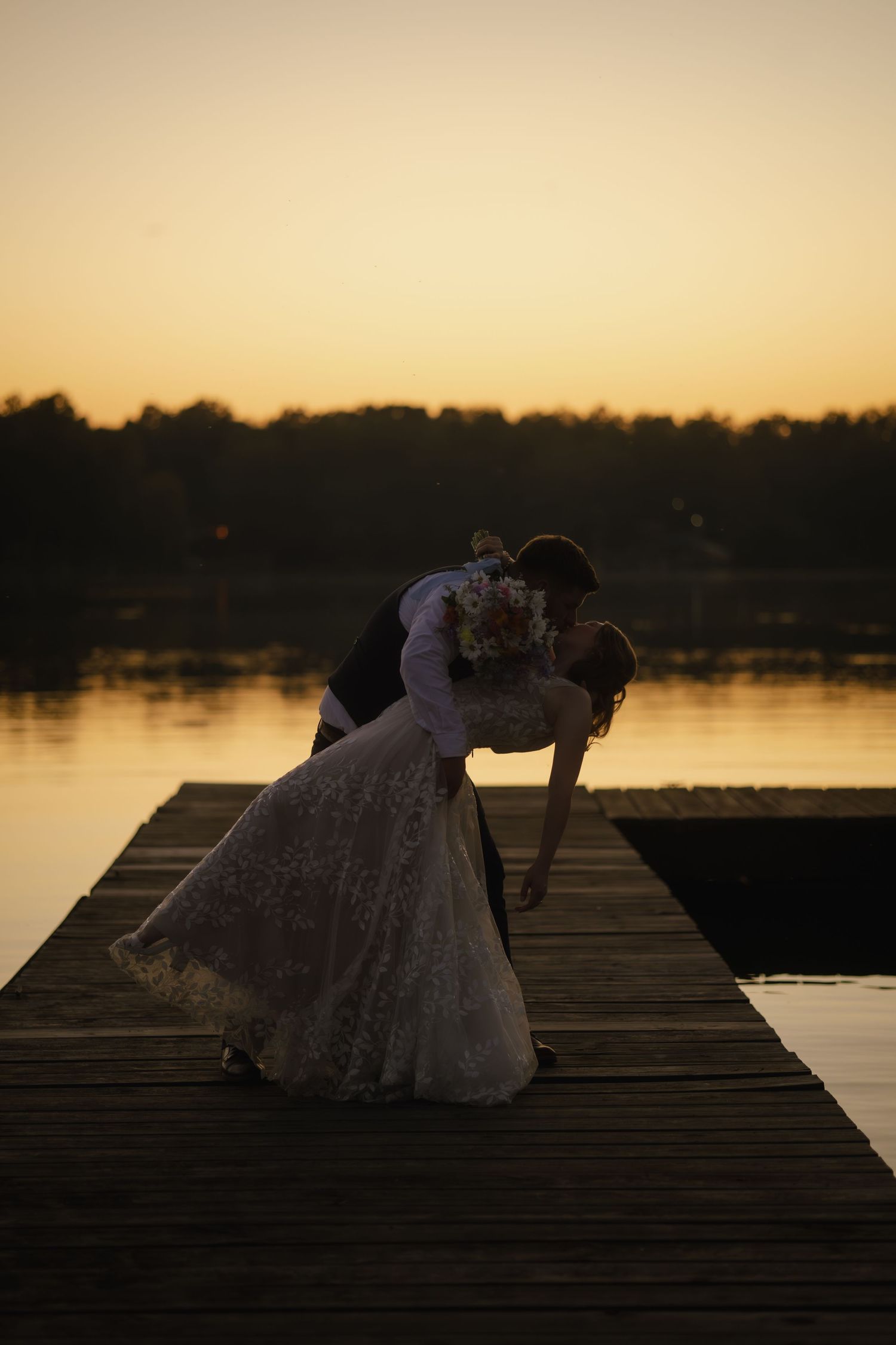 A romantic silhouette of a couple embracing on a dock at sunset with a lake in the background.