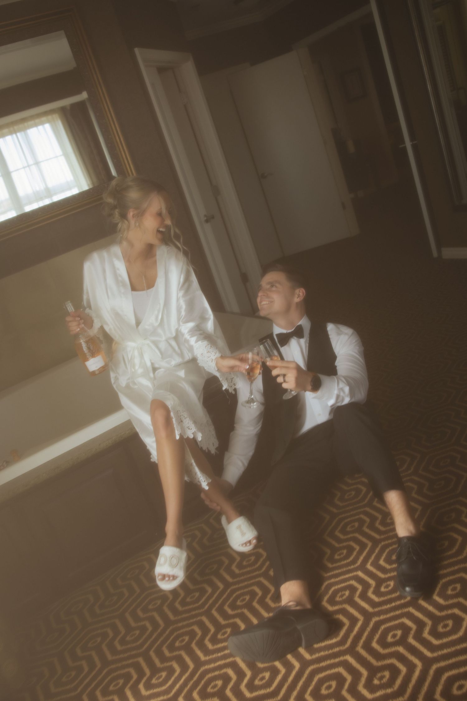 A couple in wedding attire sitting on a patterned carpet floor sharing drinks in an intimate moment.