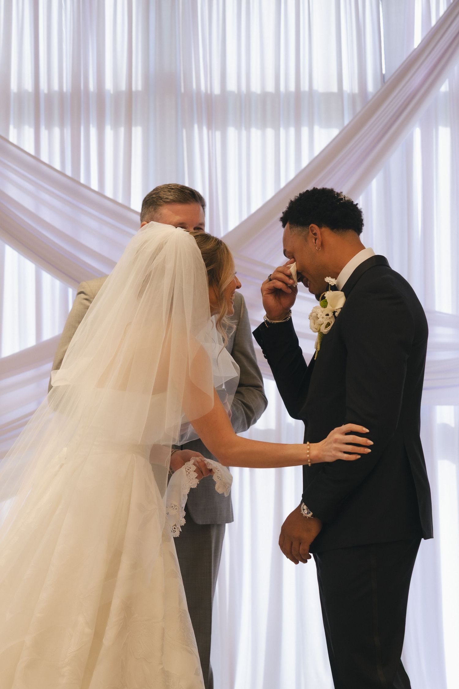 Groom kisses bride's hand at wedding altar with white draped curtains backdrop during romantic ceremony.