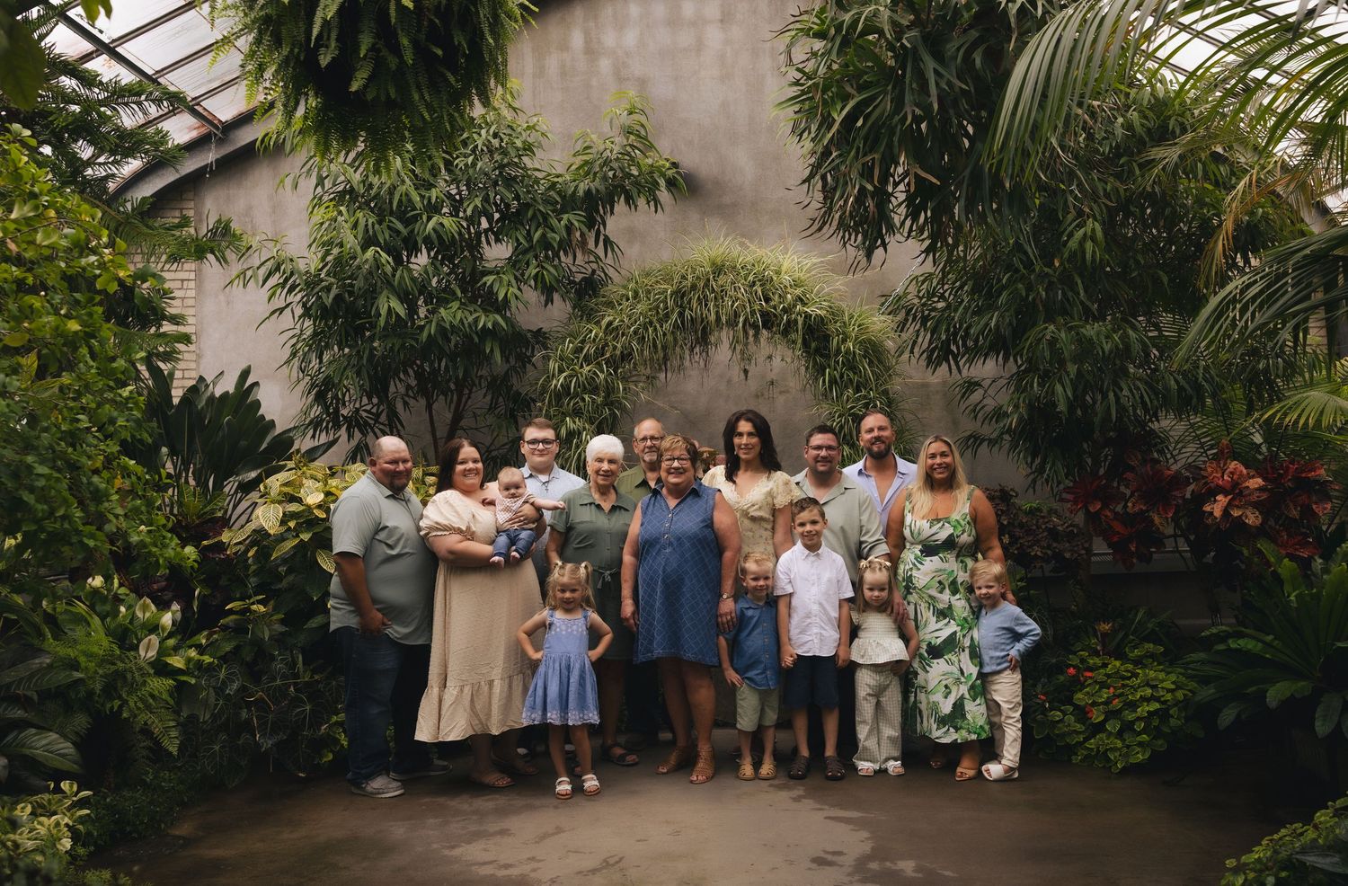Large family group portrait in a lush tropical greenhouse with green plants and flowers in the background.