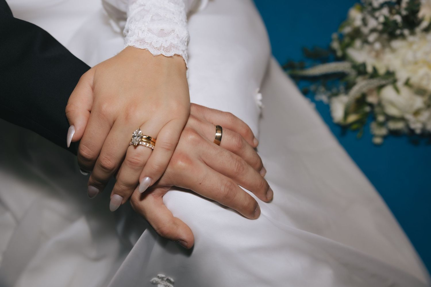 Close-up of newlywed couple hands showing gold wedding rings and diamond engagement ring on white bridal gown.