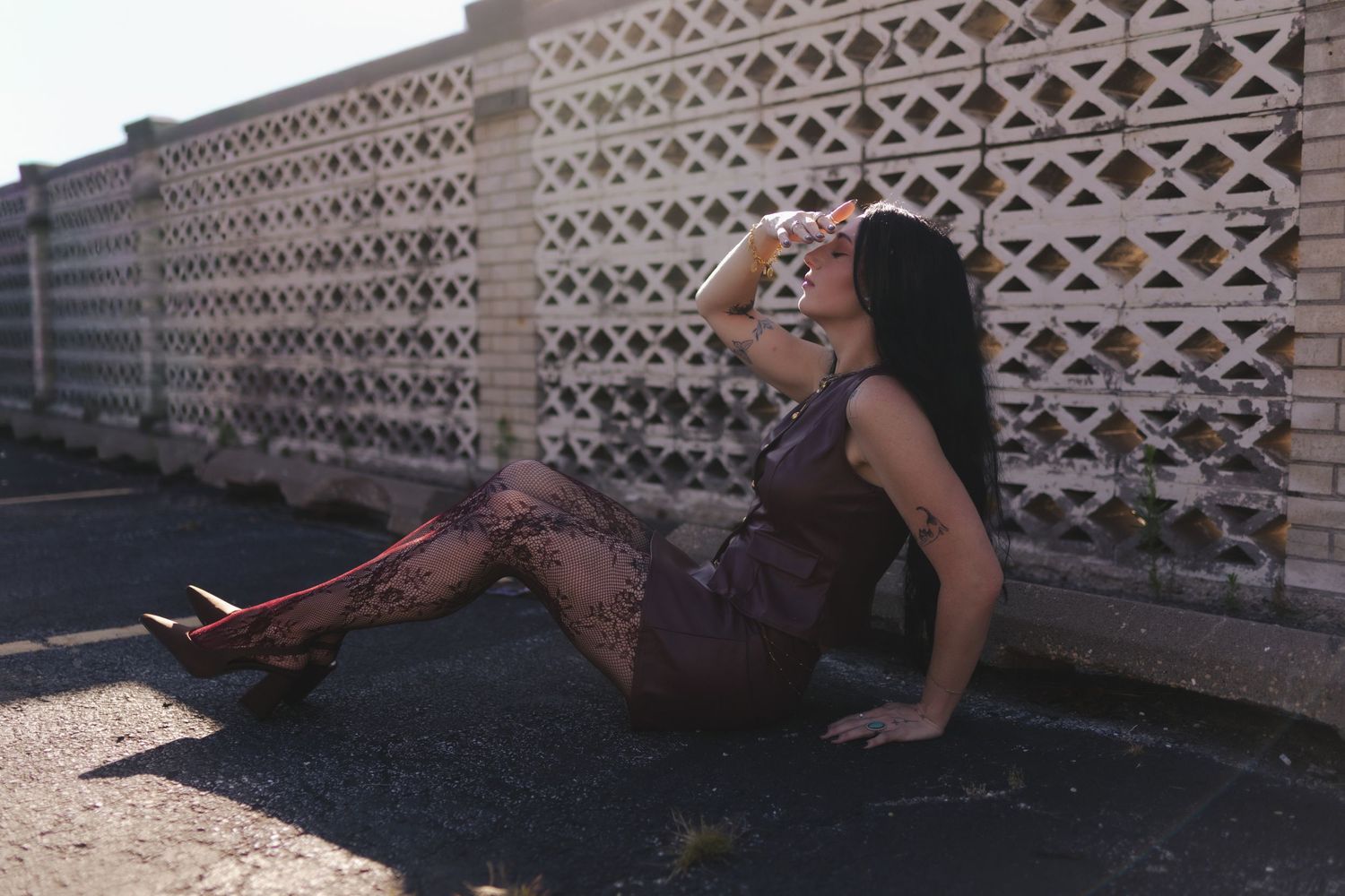 Woman in black lace dress and red heels sitting against decorative white concrete fence in sunlight.