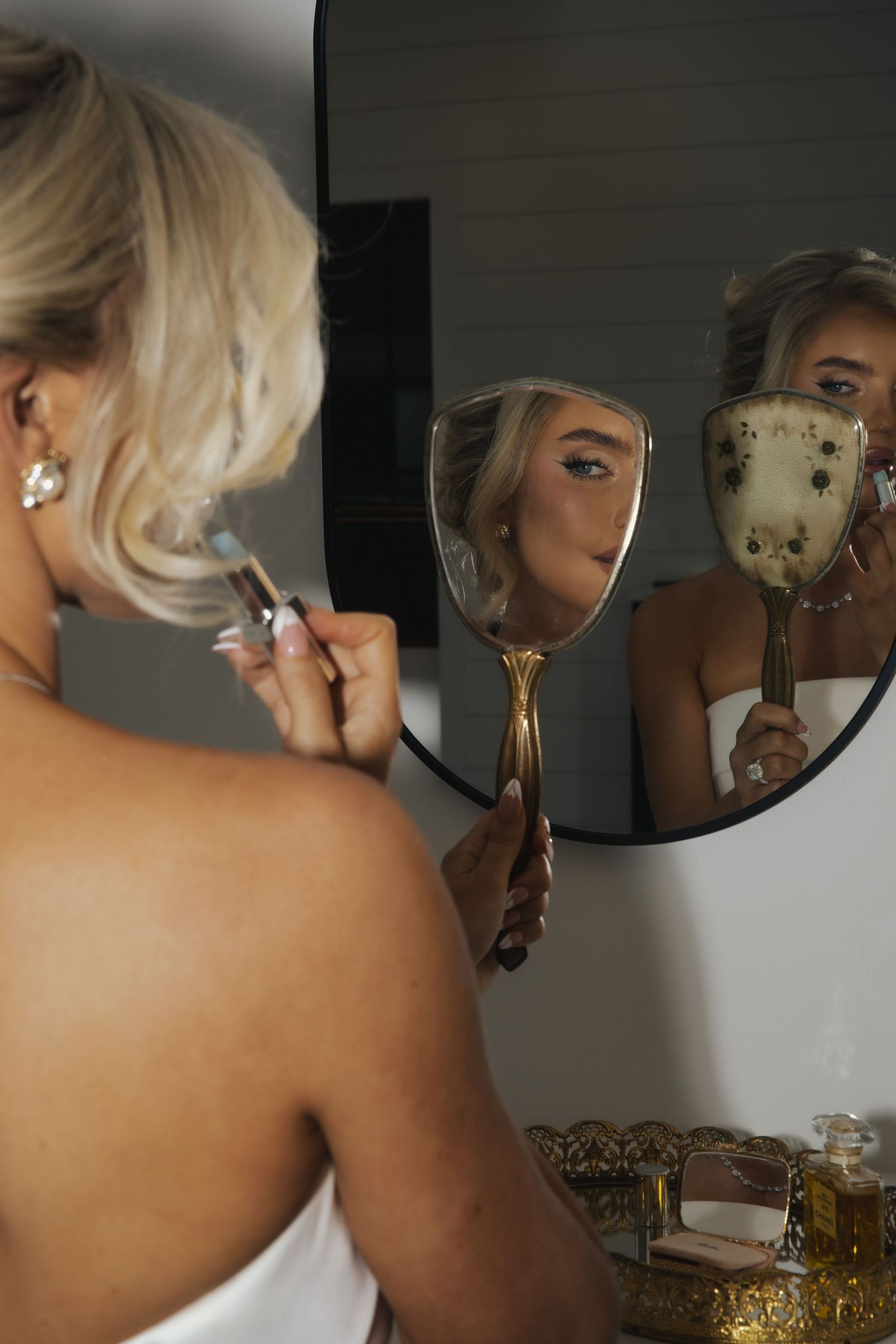 Someone applies makeup in front of a mirror while holding an ornate handheld mirror and brush in an artistic composition.