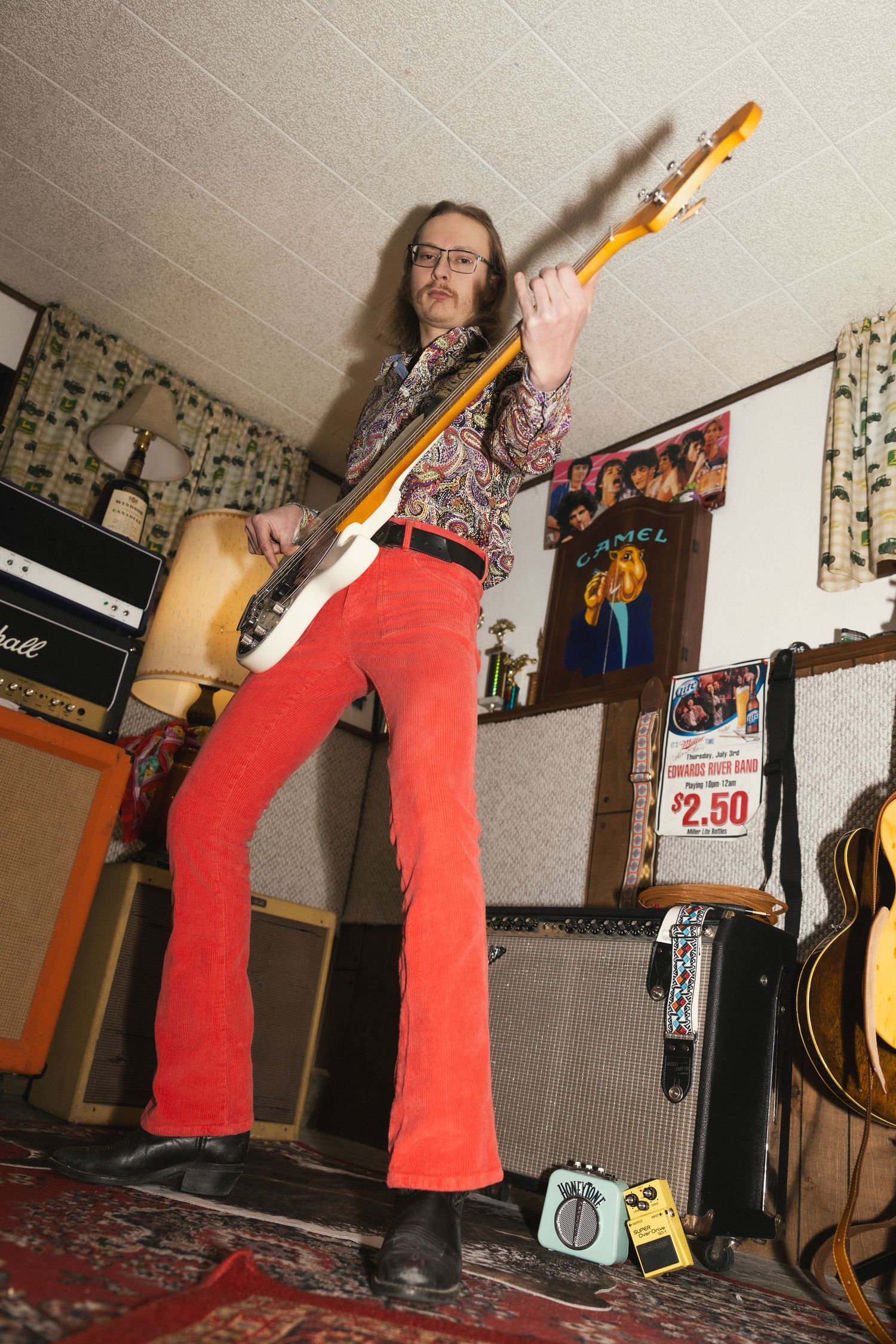 Musician in red pants plays electric guitar in a room with music equipment and floral curtains.