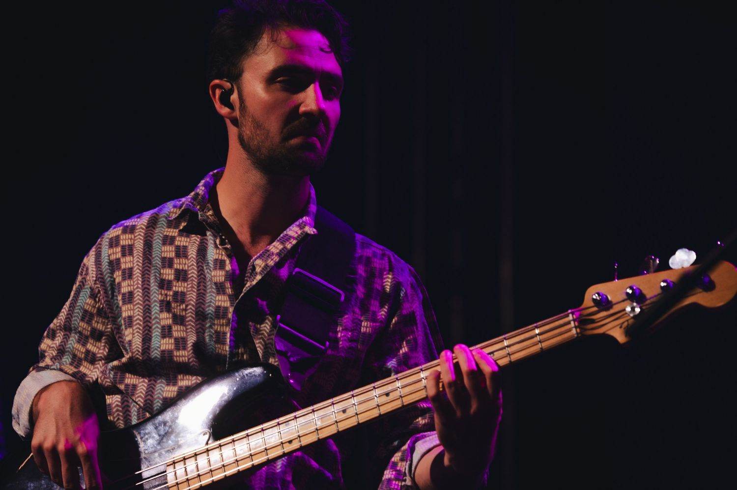 Young male musician playing bass guitar under purple stage lights at live concert.