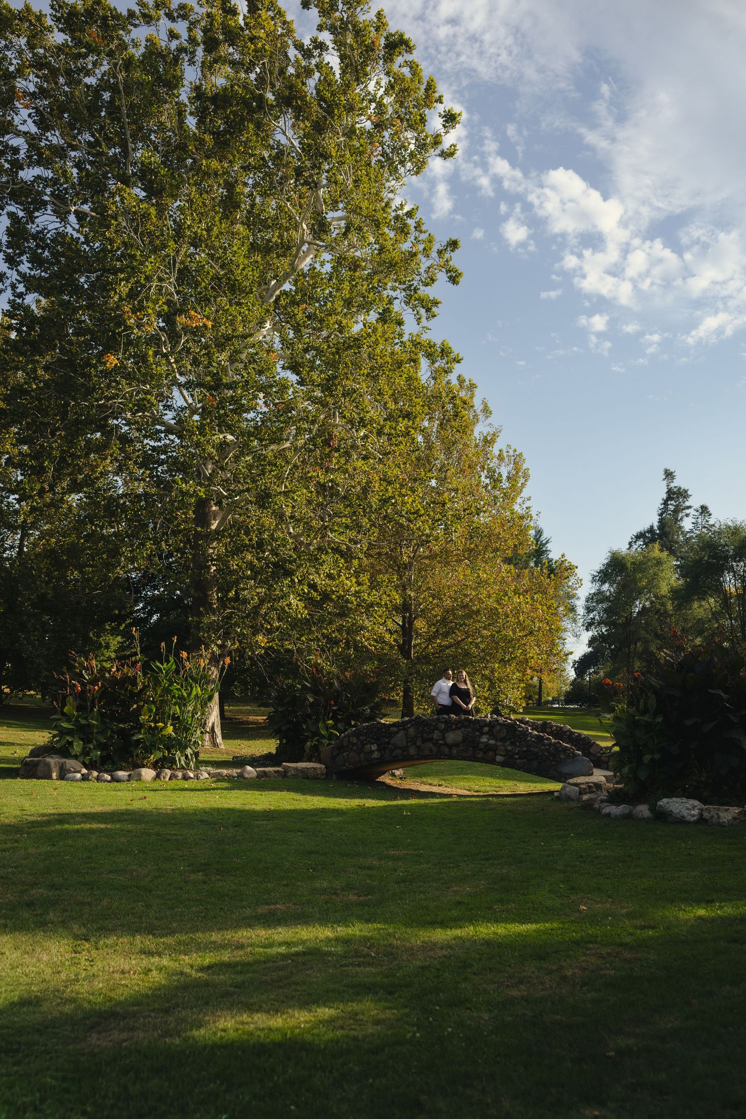 Couple sitting on stone bridge surrounded by lush green trees and manicured lawn in a sunny park.