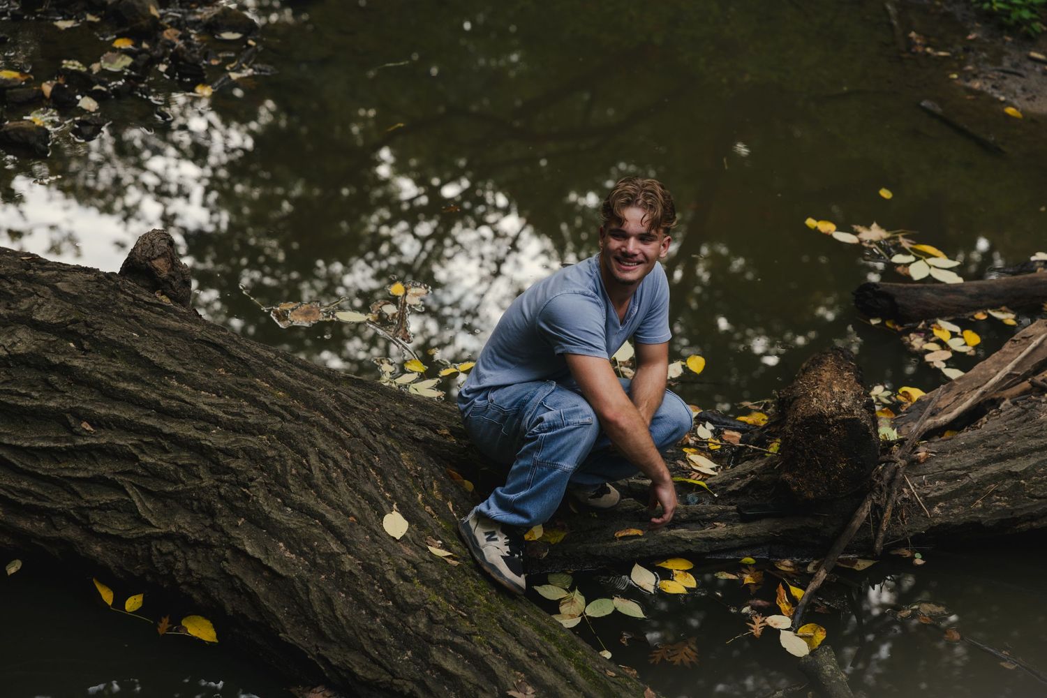 Man smiling while crouching on a fallen log over a forest pond surrounded by autumn leaves.