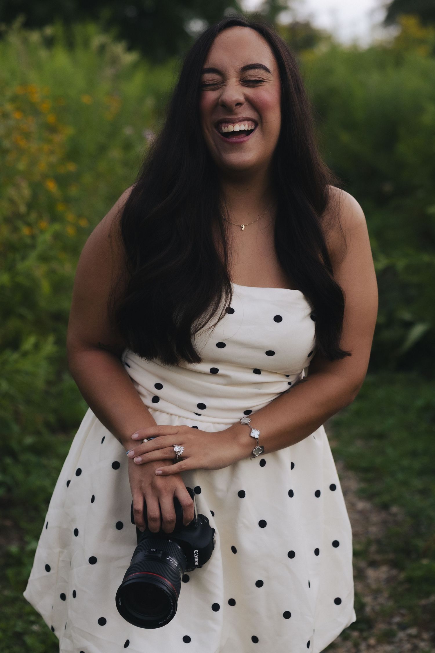 Laughing woman in white polka dot dress holding a Canon camera outdoors in a lush green garden setting.
