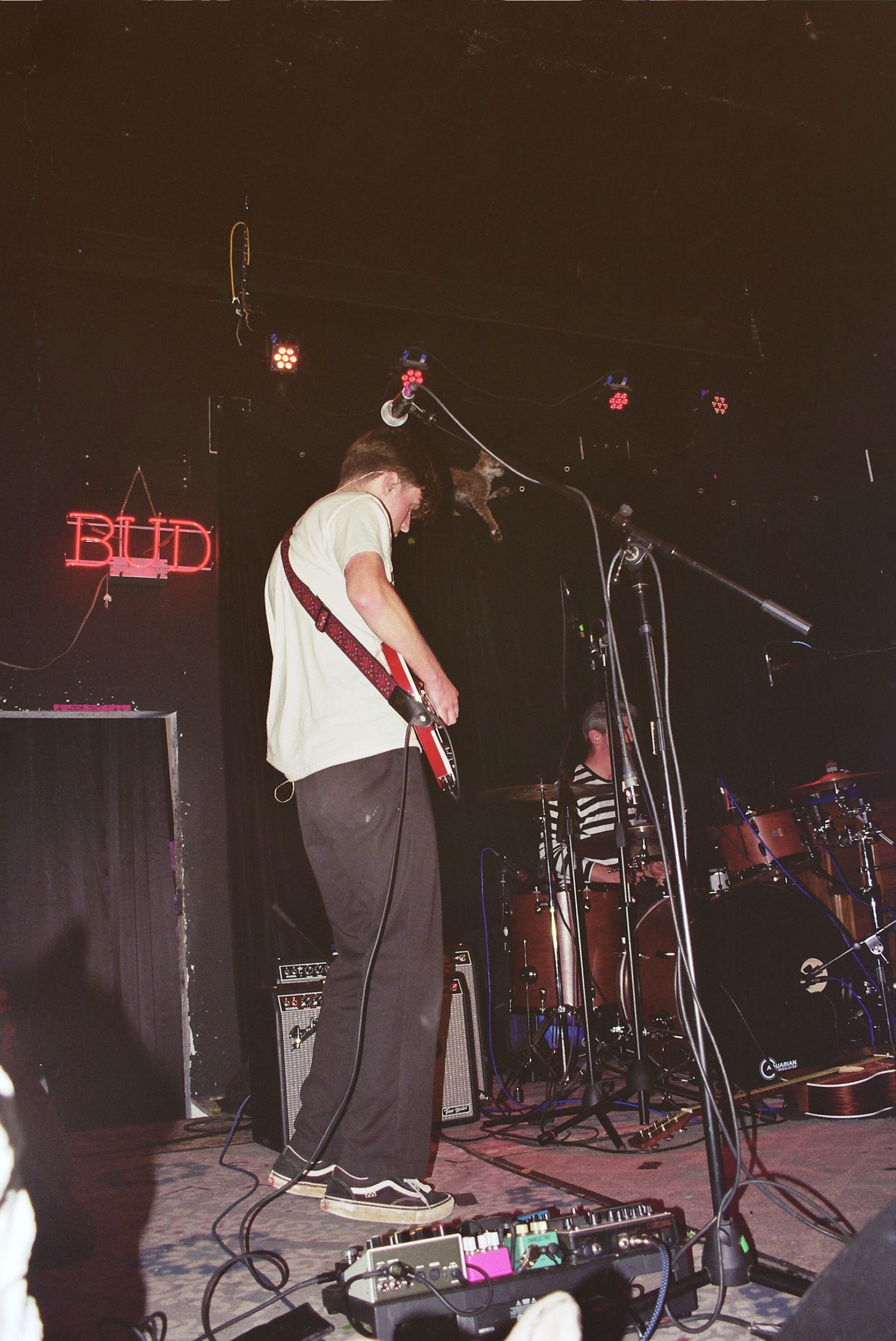 Guitarist performing on stage at a live music venue with a Budweiser neon sign and drum kit visible in the background.