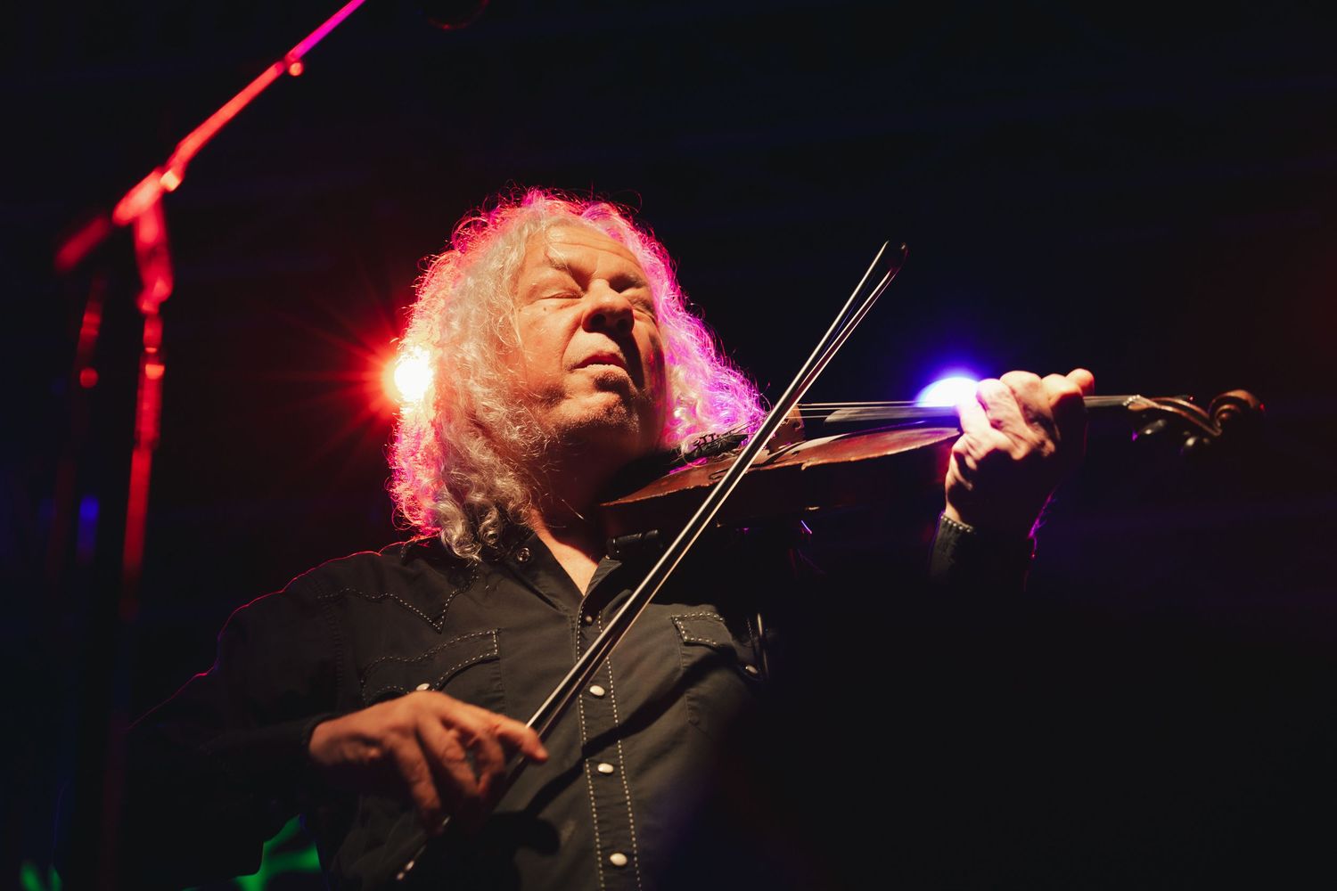 A curly-haired musician plays violin on a dimly lit stage with colorful lights behind him.