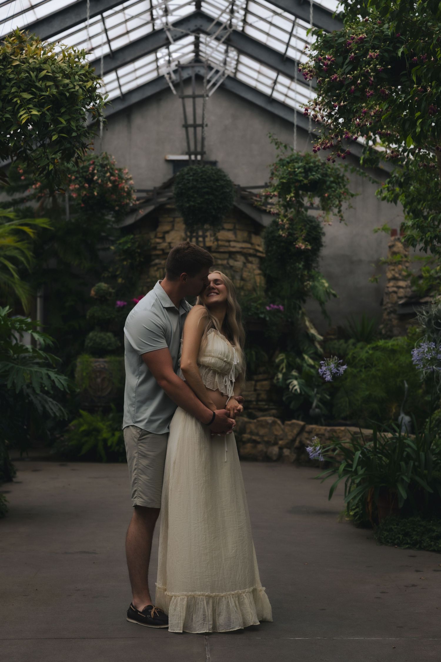 Couple embracing in a lush greenhouse, woman in white dress smiling as man leans in close.