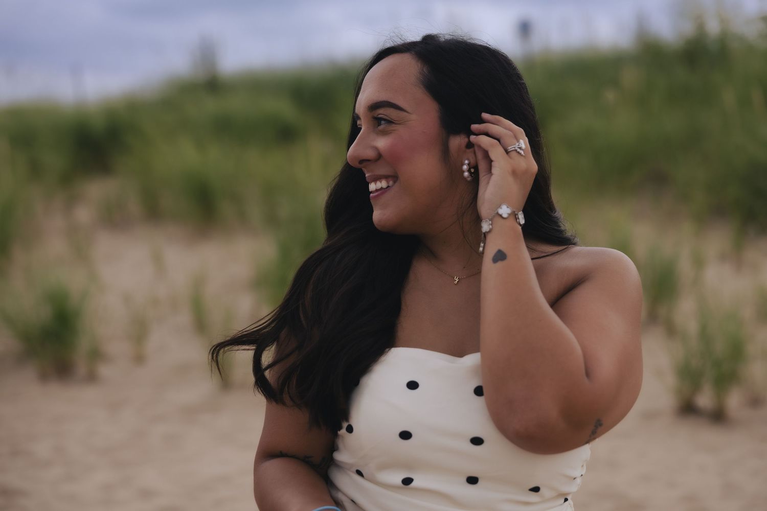 A smiling woman with long dark hair in a white polka dot top touches her ear on a sandy beach.