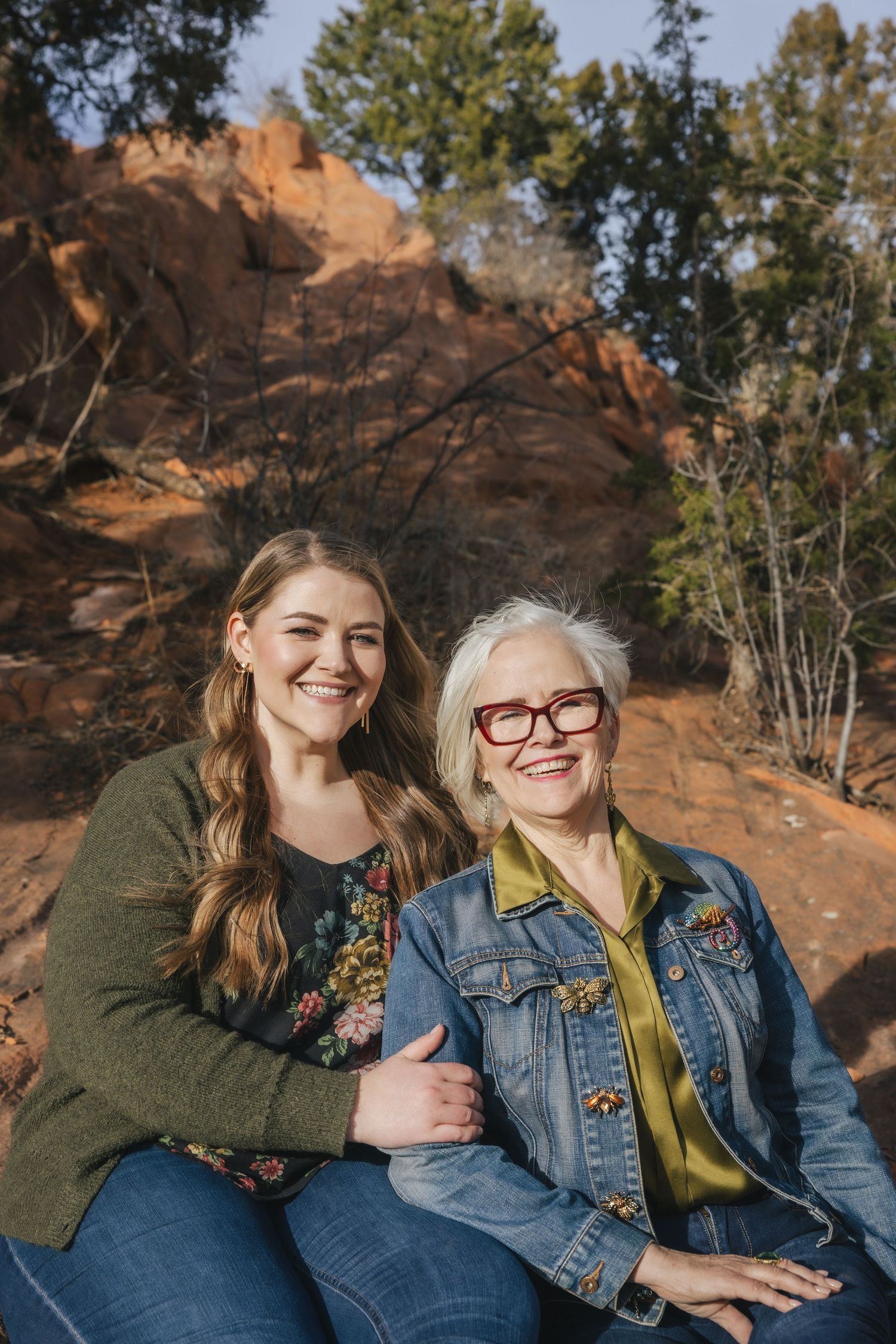 Two women smiling outdoors near red rock formations and trees, posing together for a portrait photo.