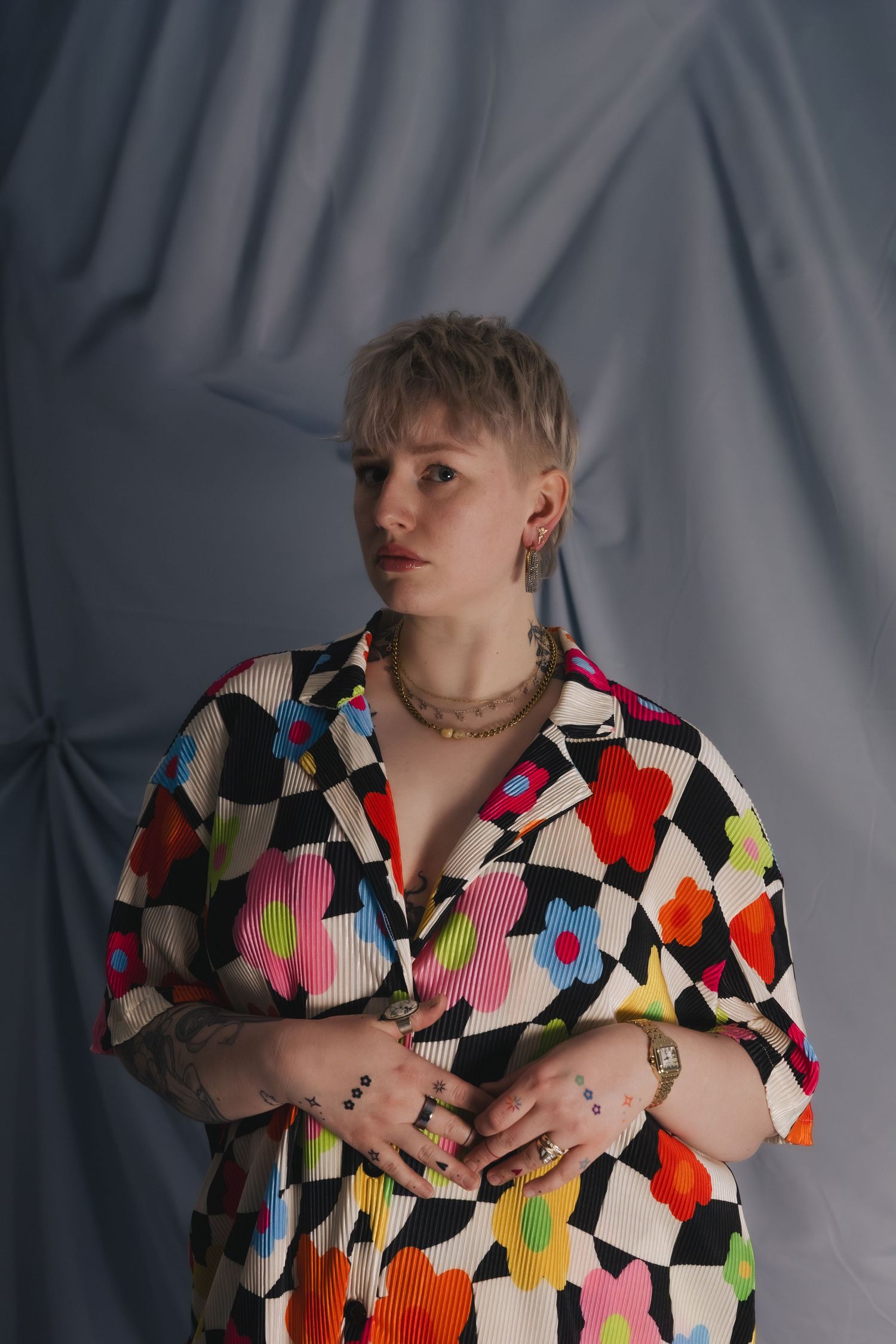 Person with short blonde hair wearing a colorful floral checkered shirt, gold jewelry, and rings, posing against a gray backdrop.