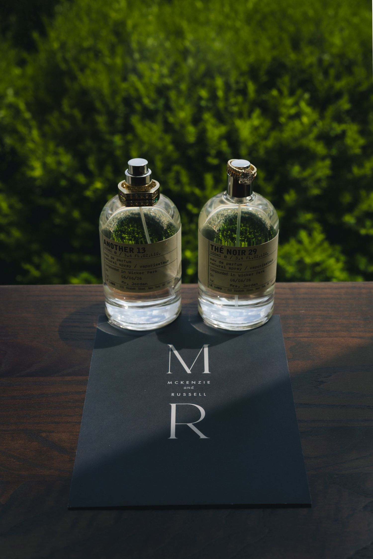 Two glass fragrance bottles on a wooden surface beside a dark McKenzie Russell branded card, with green foliage background.