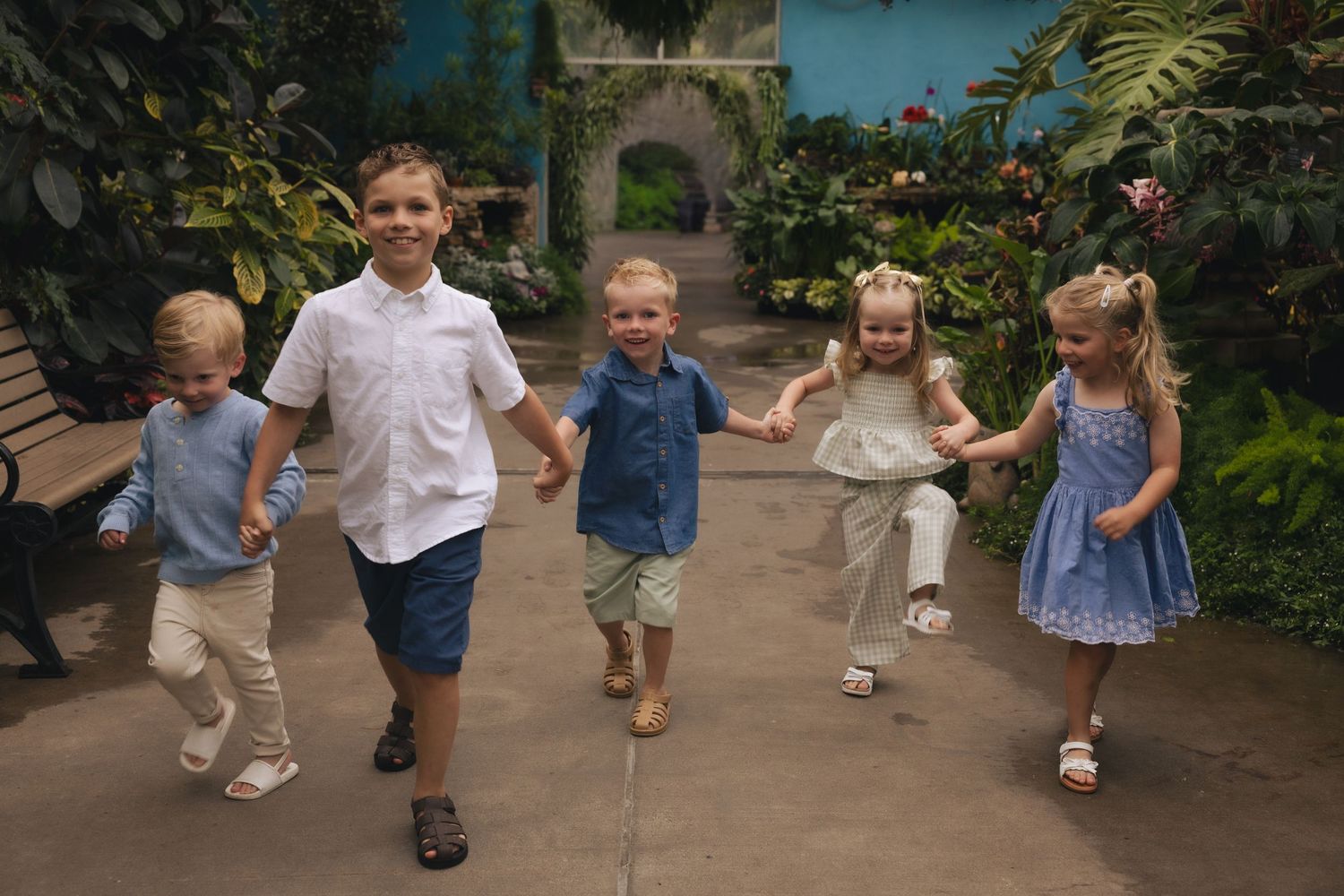 Five young children holding hands and running together through a lush indoor garden with tropical plants.