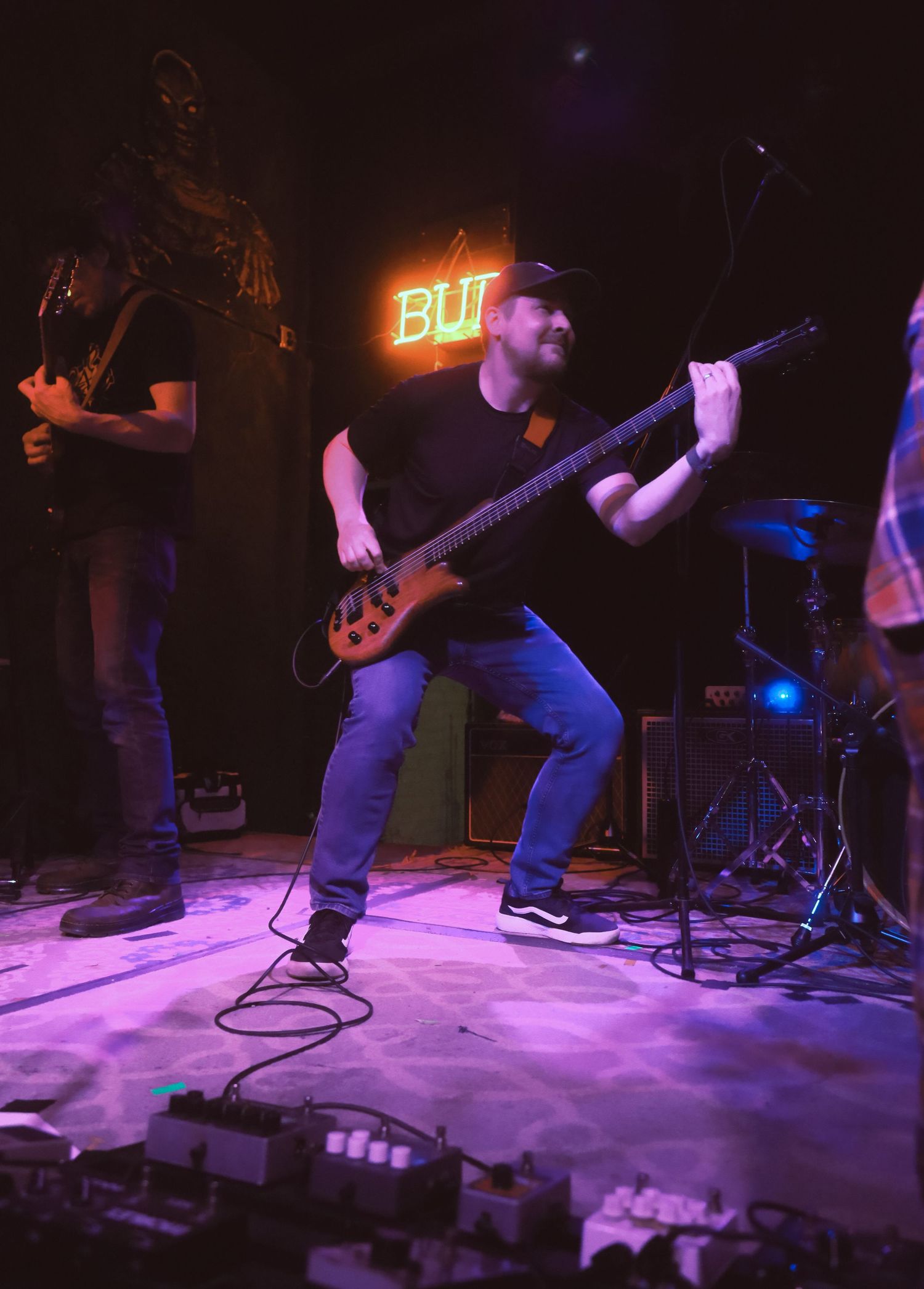Musicians perform on stage with guitar and neon 'BUT' sign glowing in purple and blue lighting.