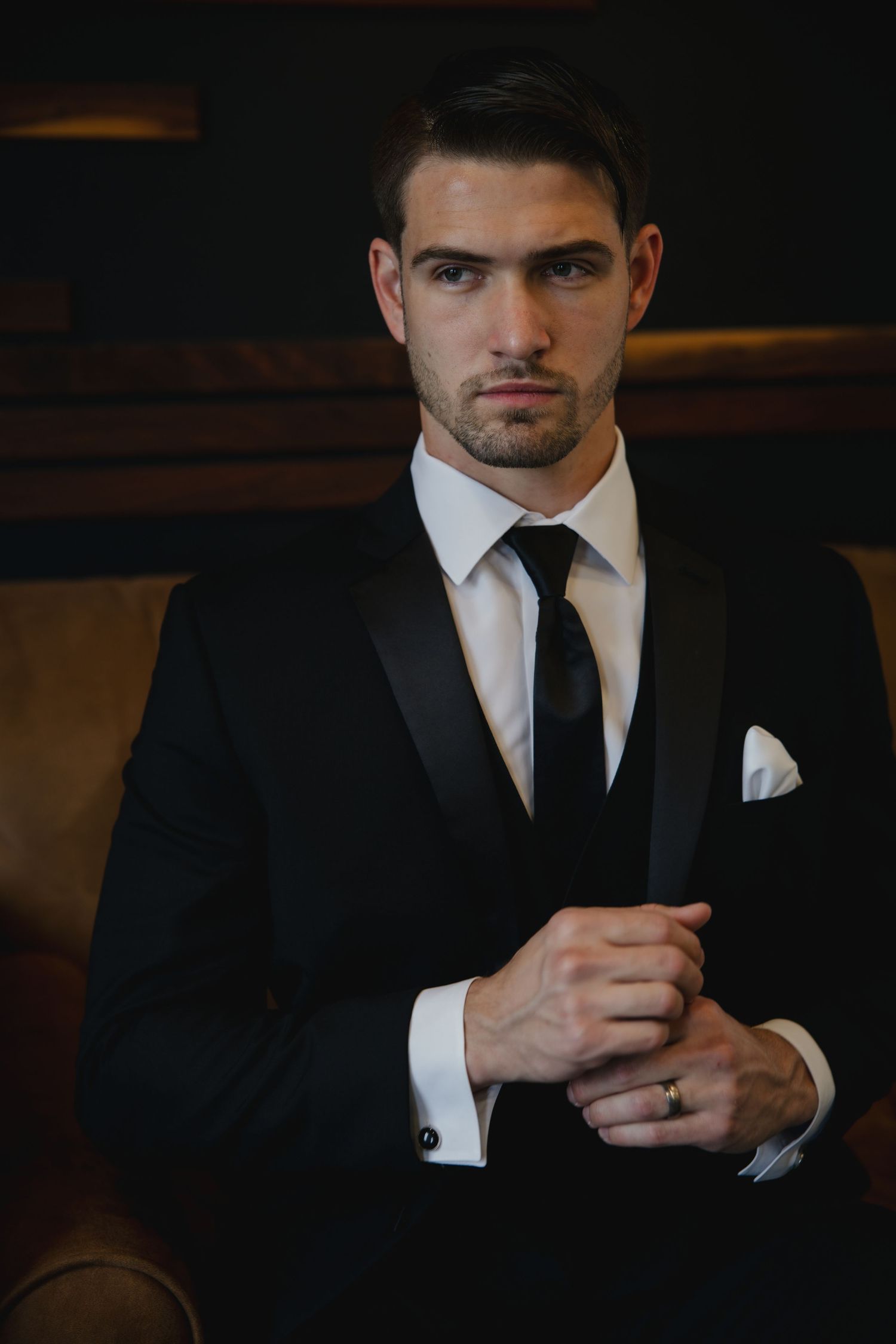 Dark and moody portrait series showing a person in formal black suit with white shirt and black tie against dark background.