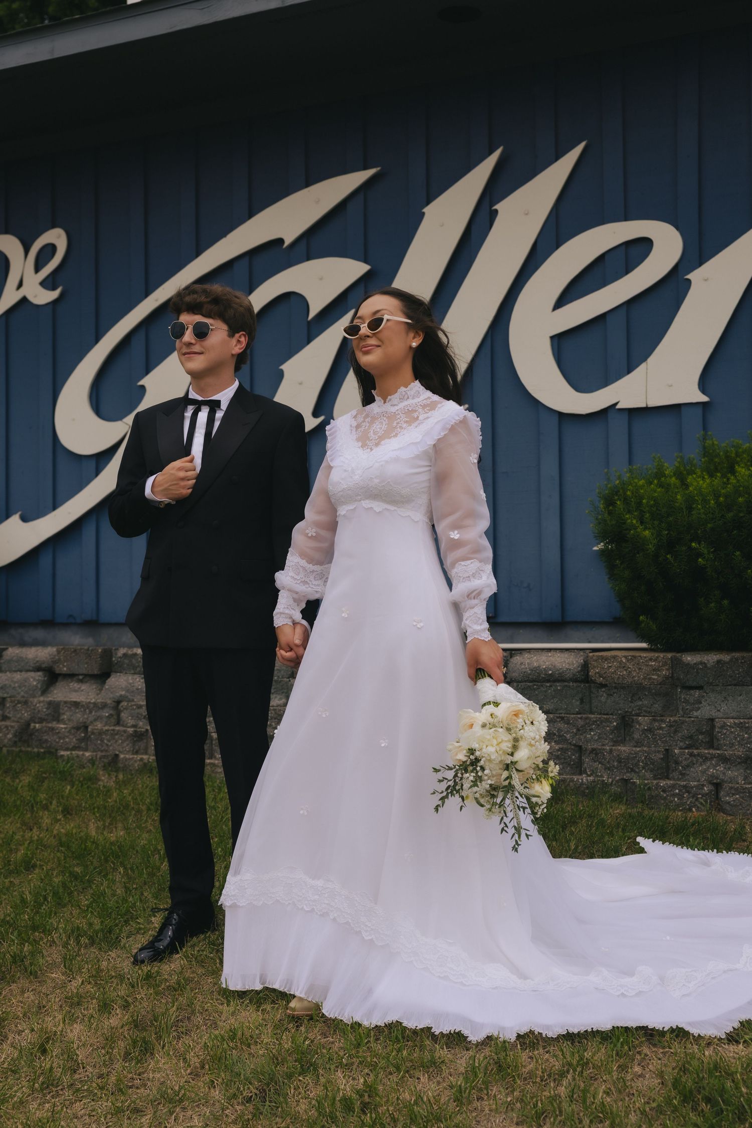 A bride in a white gown holding a bouquet and groom in black suit pose holding hands outside a blue building.