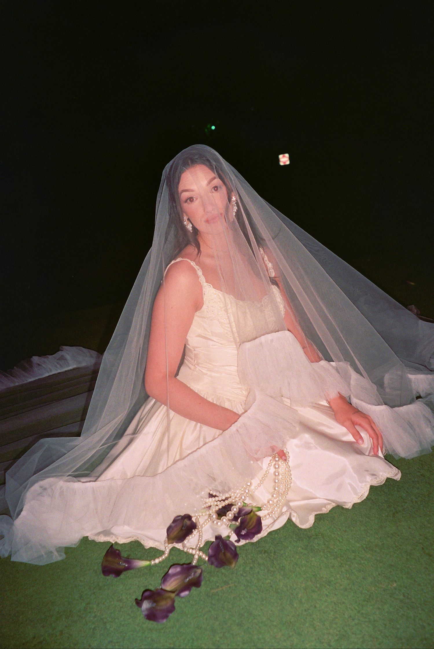 Bride in white dress and veil sits on grass at night, surrounded by purple rose petals and pearl accessories.