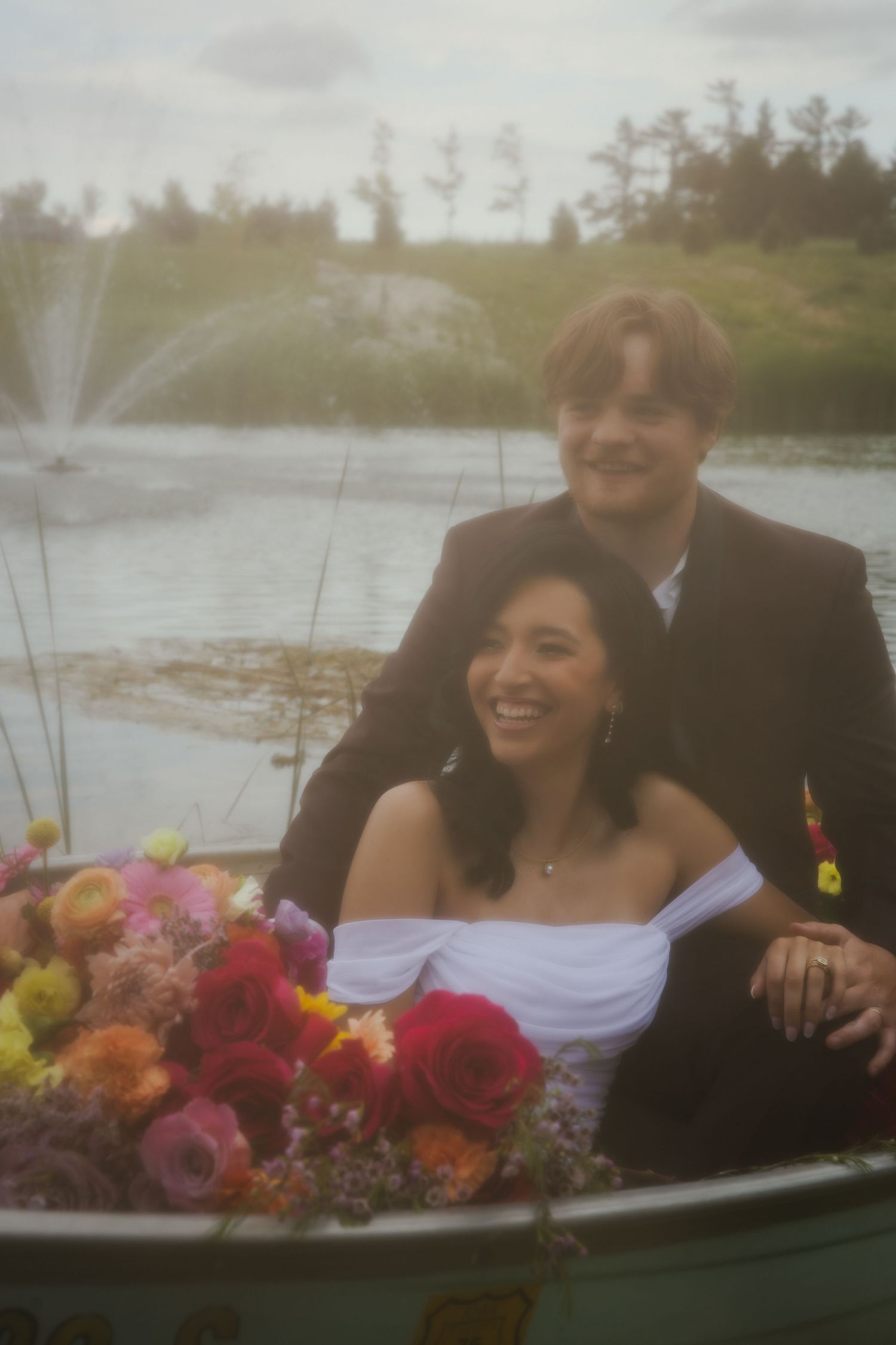 A couple poses with vibrant floral bouquets near water on their wedding day, laughing and sharing joyful moments together.